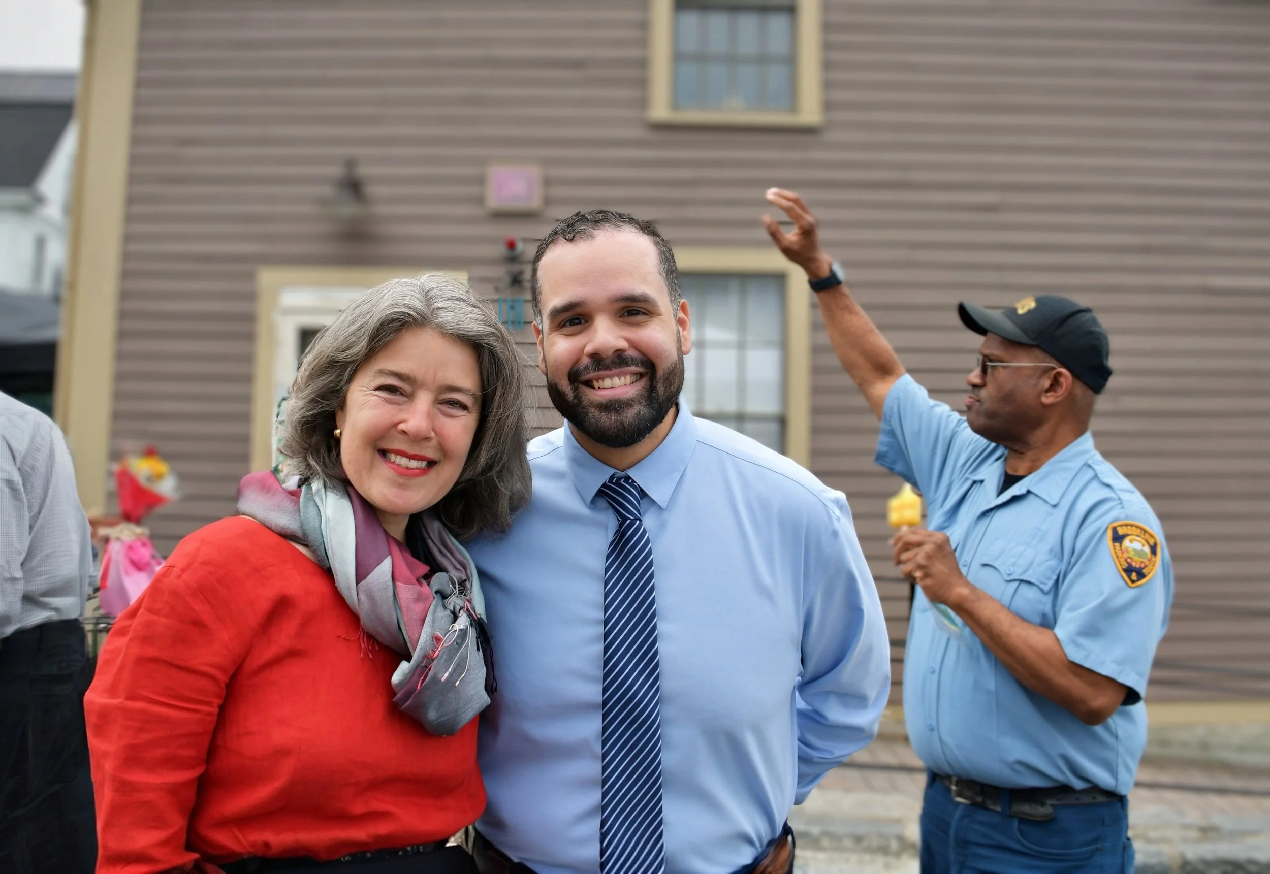 A group of people smiling outdoors, with a woman wearing a red jacket and scarf, and a man in a blue shirt and tie in the foreground. Two other men in uniform are in the background.