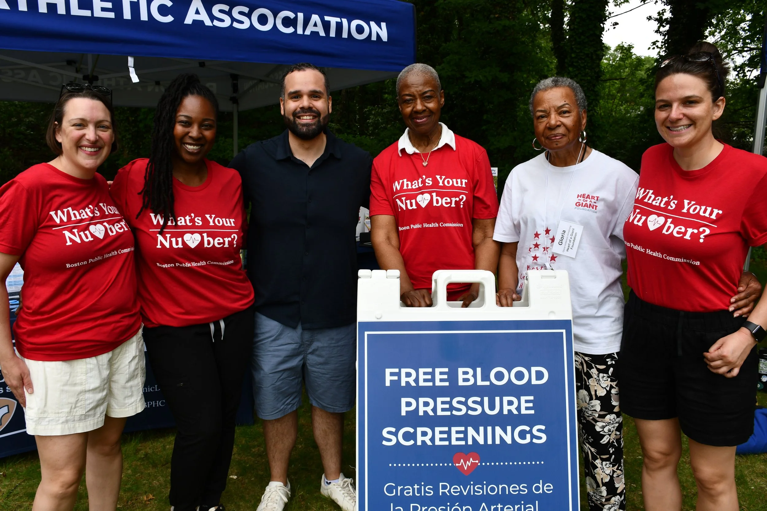 Group of six diverse people standing behind a table at an outdoor health event, smiling, some wearing red shirts with health-related slogans, with a blue canopy above them. The sign indicates free blood pressure screenings.