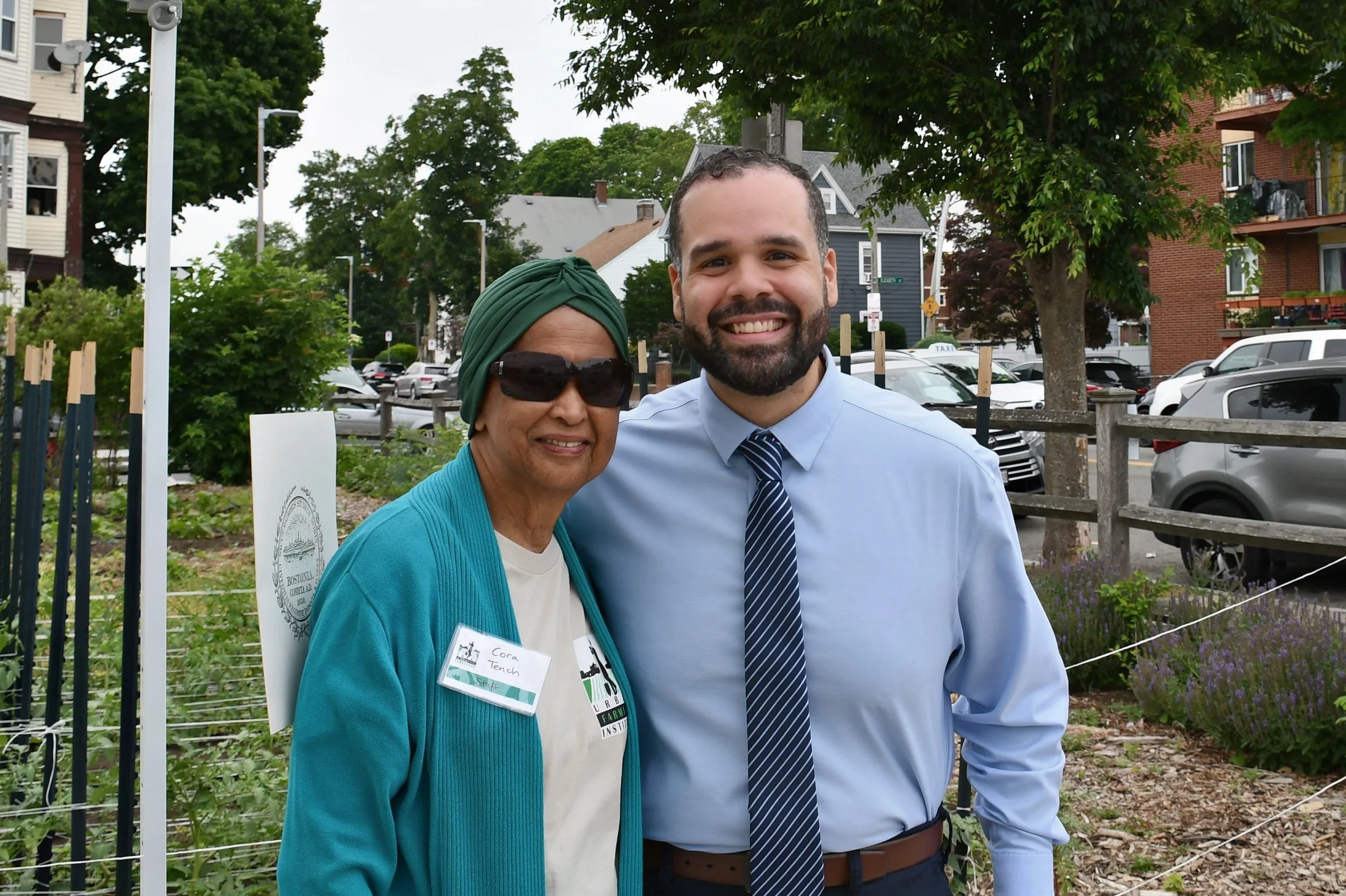 A woman with a green head wrap, sunglasses, and a teal cardigan standing next to a man with short dark hair, beard, wearing a light blue dress shirt, striped tie, smiling outdoors in front of trees, parked cars, and a brick building.