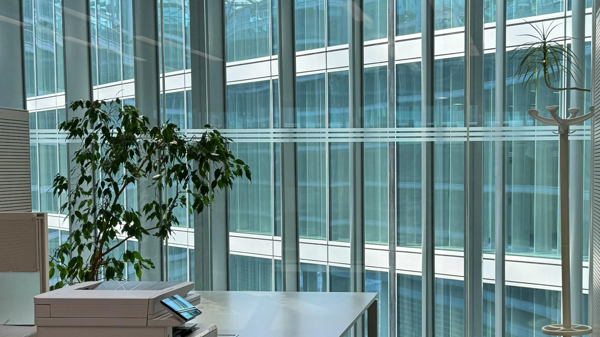 Interior of a modern office building with large floor-to-ceiling glass windows, plants, and a small digital device on a white desk.