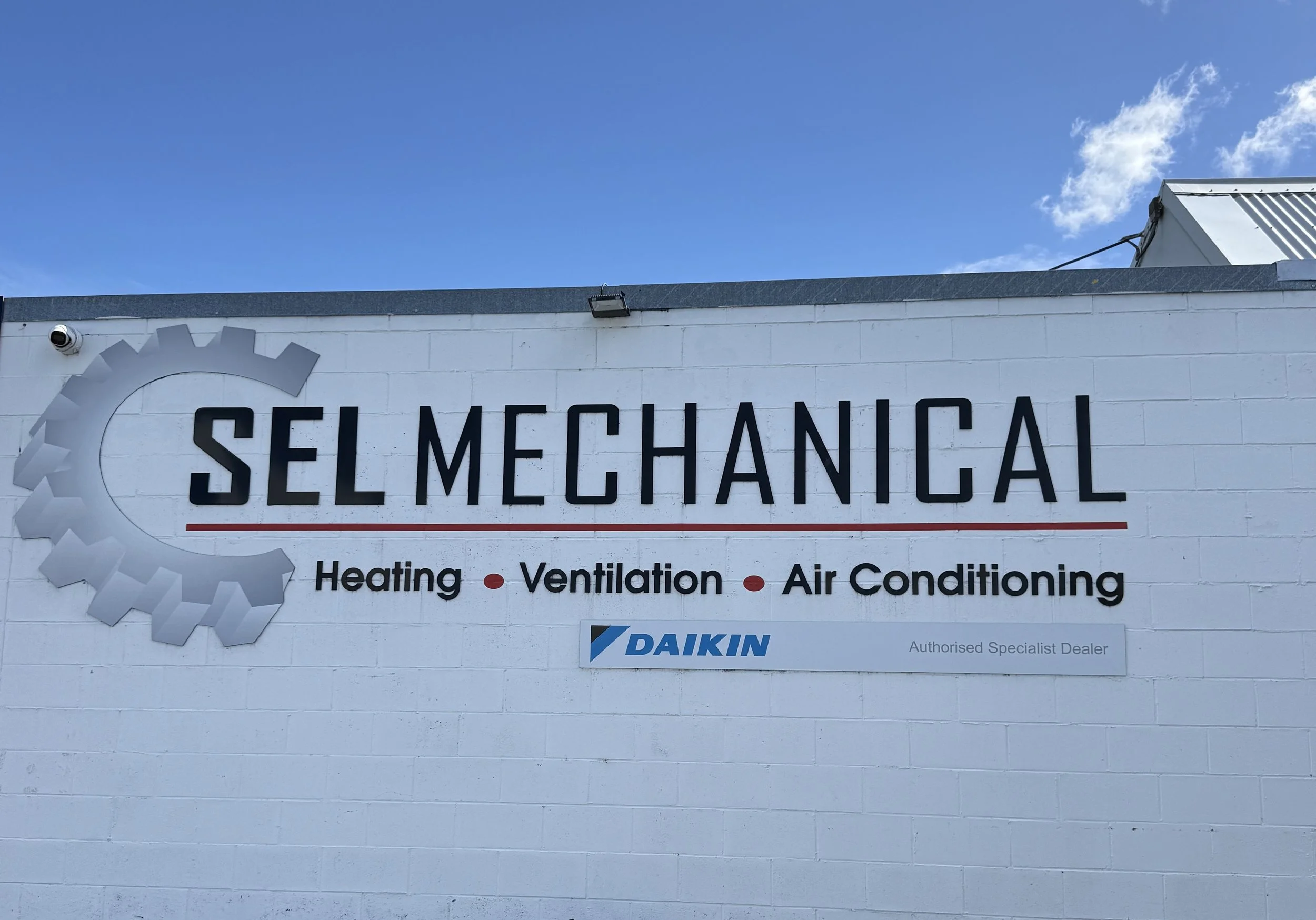 A sign on a white brick building displays the company name "SEL Mechanical" with a gear logo. The sign lists services: Heating, Ventilation, and Air Conditioning. The background shows a blue sky with a few clouds.