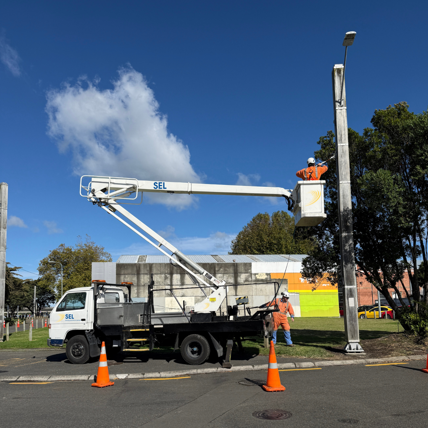 Workers using a cherry picker lift on a street to install or maintain a streetlight or utility pole, with traffic cones around the work area, trees and buildings in the background, and a partly cloudy sky.