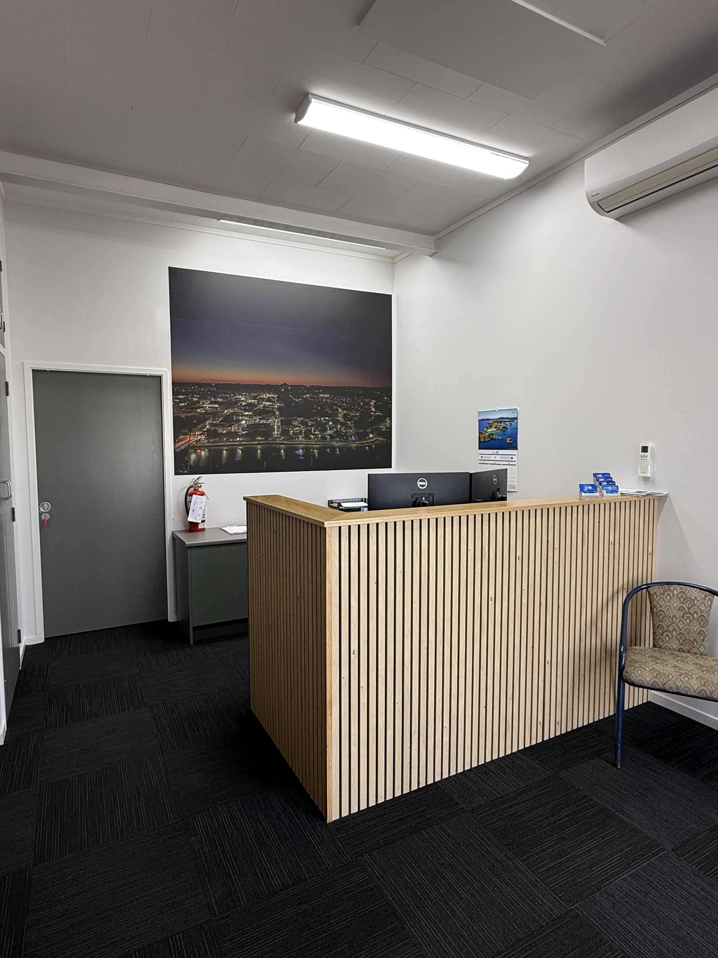 The image shows a reception desk made of light-colored vertical wooden slats. Behind the desk are two computer monitors. To the right of the desk, a patterned chair with metal legs is visible. Large black and white framed landscape photograph of a cityscape at sunset hangs on the wall. A gray cabinet with a fire extinguisher on top is next to a door with a frosted glass panel. The room has a white ceiling with two fluorescent light fixtures and an air conditioning unit. The floor is carpeted with black textured tiles.