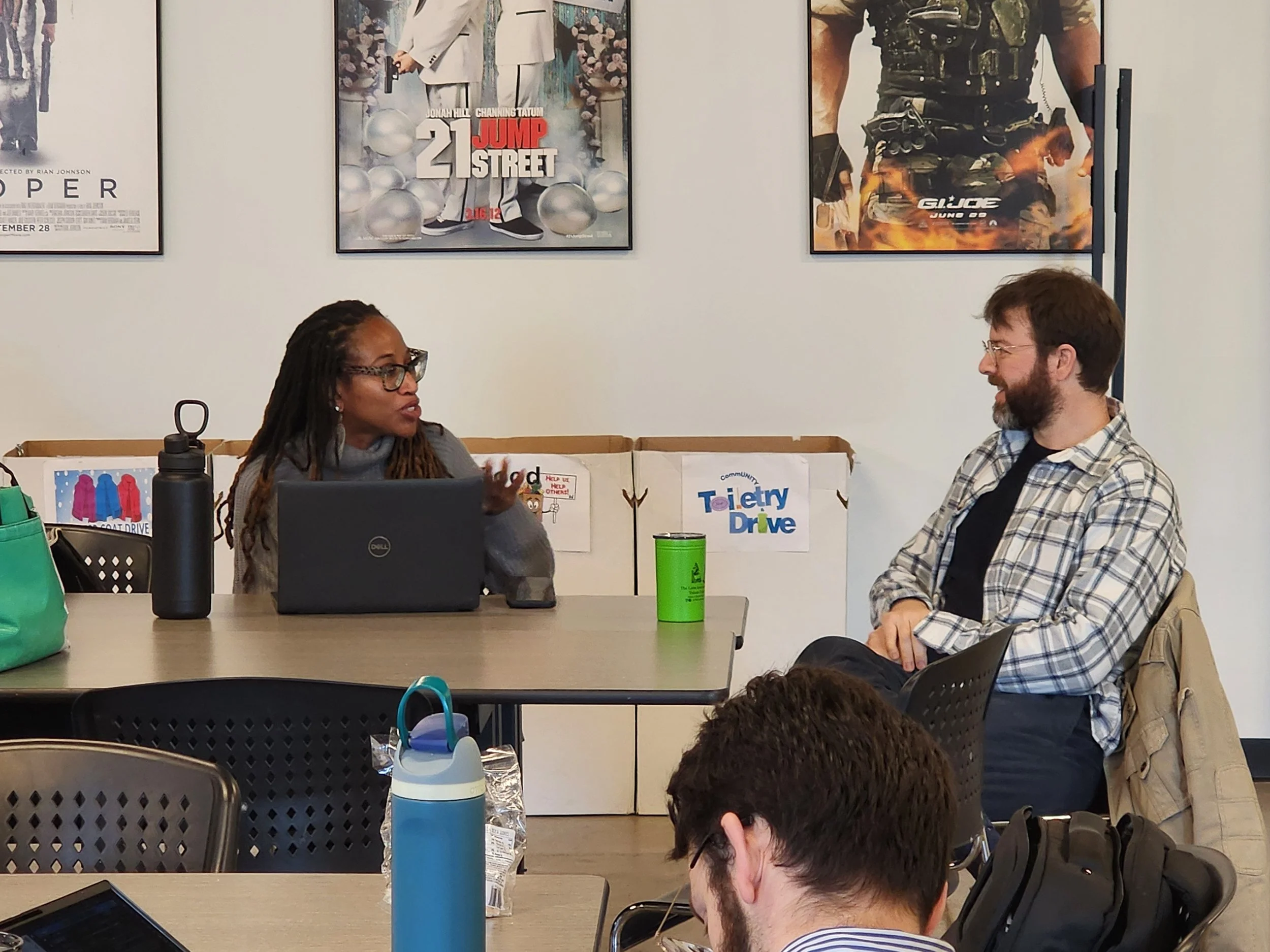 Two people sitting at a table in a meeting room, engaged in conversation. The woman on the left has dreadlocks and glasses, and the man on the right has brown hair, a beard, and glasses. The background features posters on the wall and a sign that says 'Community Toy Drive'.