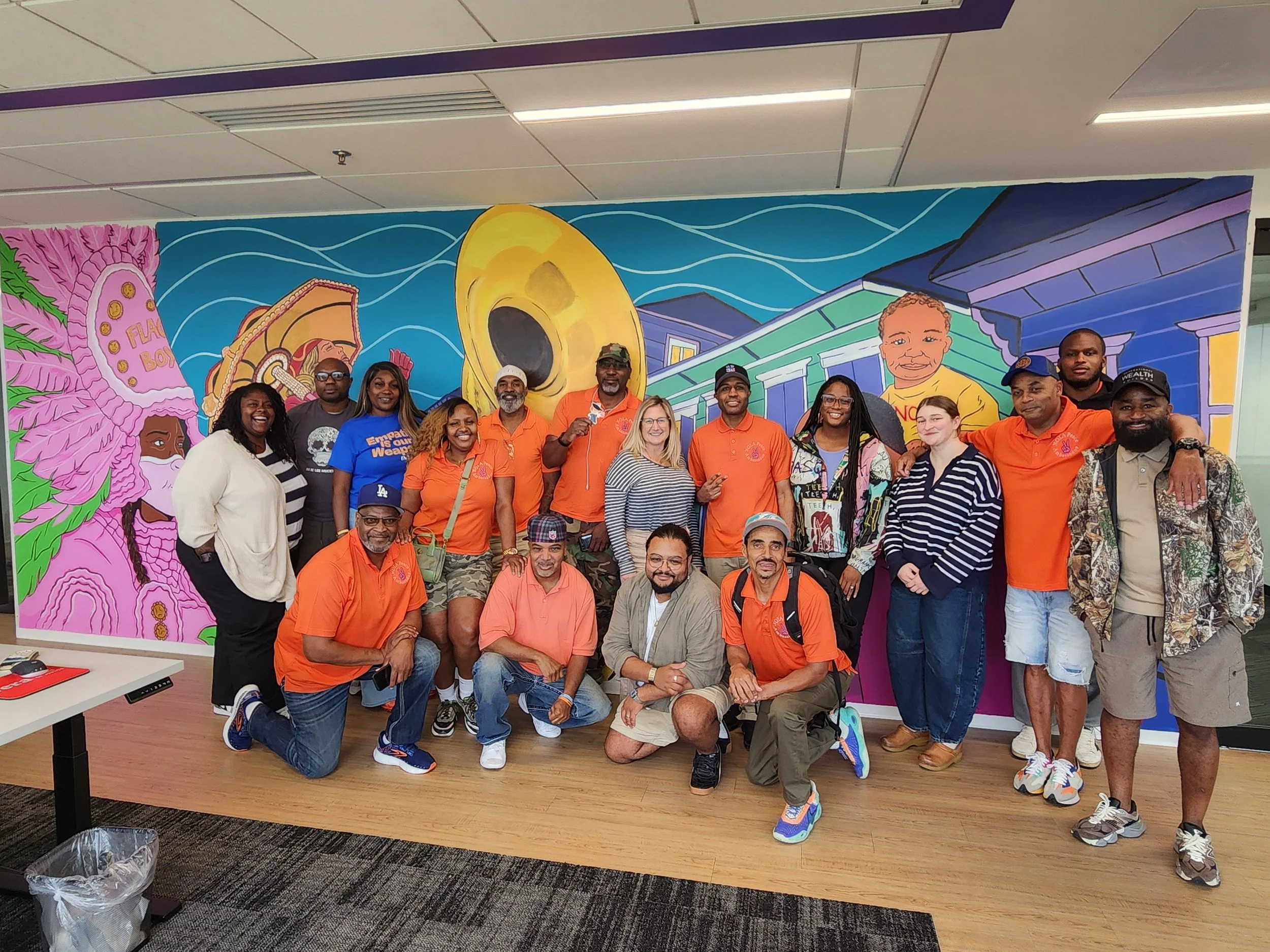 A group of diverse people gathered in front of a colorful mural inside a building. The mural features bright pink flowers, a large yellow eye, and stylized buildings. The group is smiling and wearing casual clothing, with some in orange shirts.