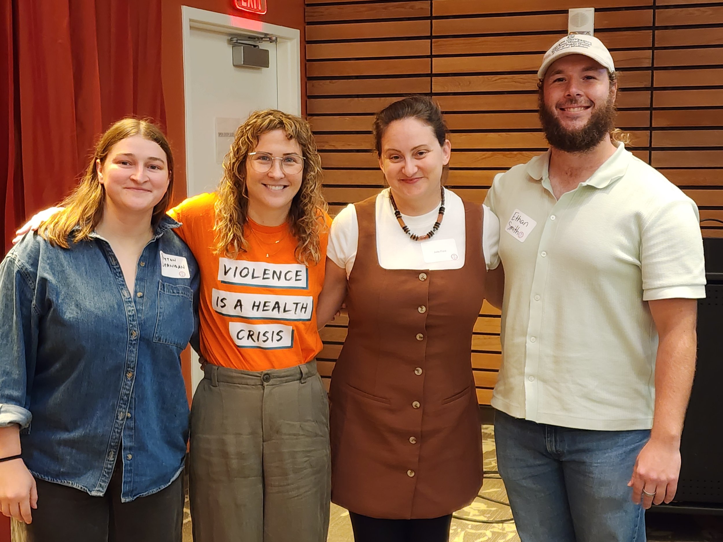 Group of four people, three women and one man, standing together indoors in front of wooden panel walls, smiling at the camera. One woman is wearing glasses, another has a shirt with a message about violence being a health crisis, and the man is wearing a white cap. They have name tags on their clothes.