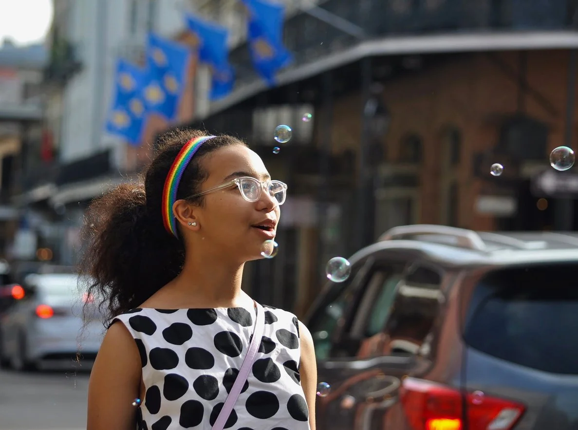 A young woman with glasses, a rainbow headband, and a black and white polka dot dress standing outdoors in a city street, with soap bubbles floating around her.