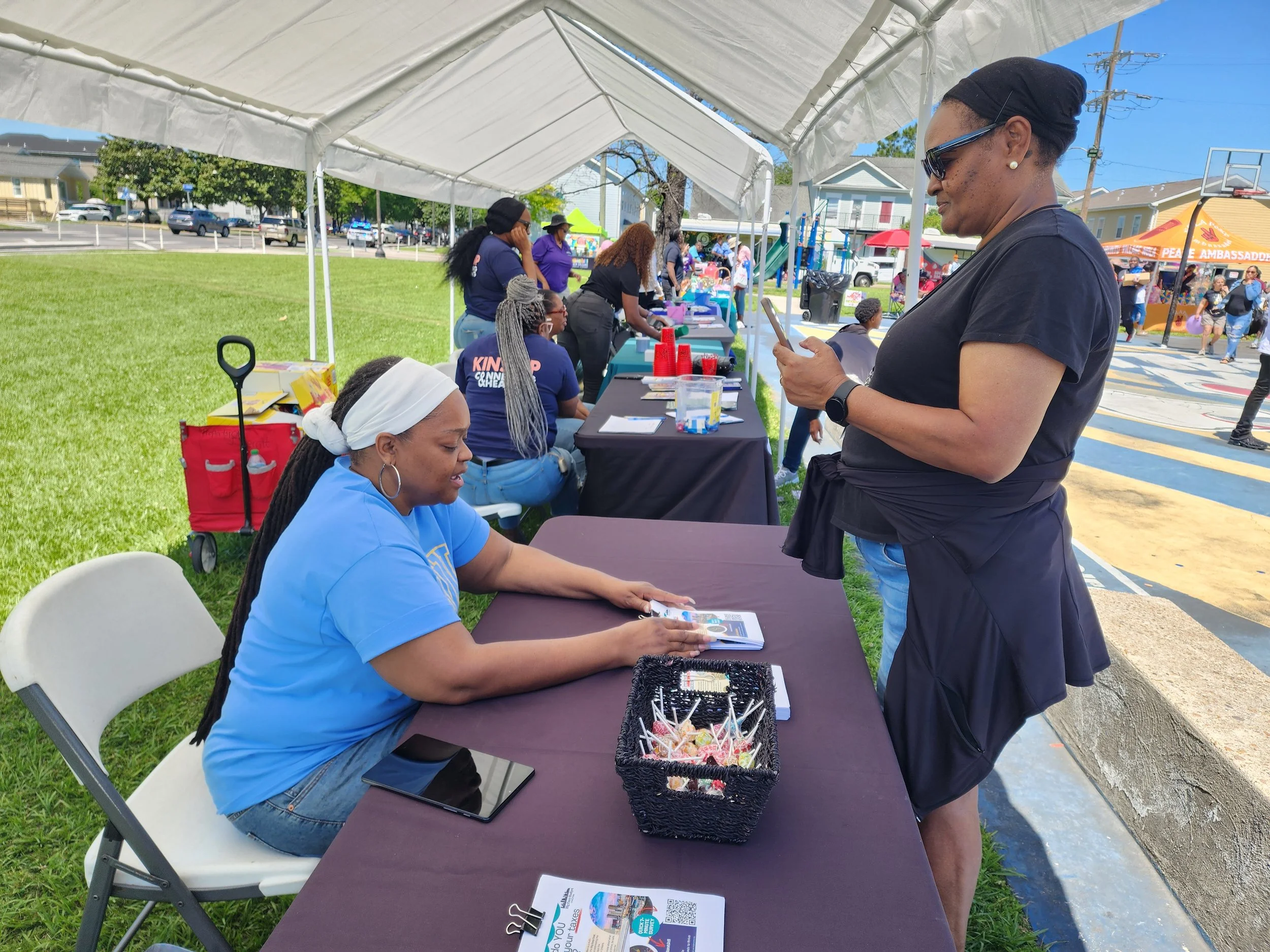 People at an outdoor event under a white canopy tent, with tables, chairs, and booths in background, some individuals seated at the table and one person standing and looking at a smartphone.