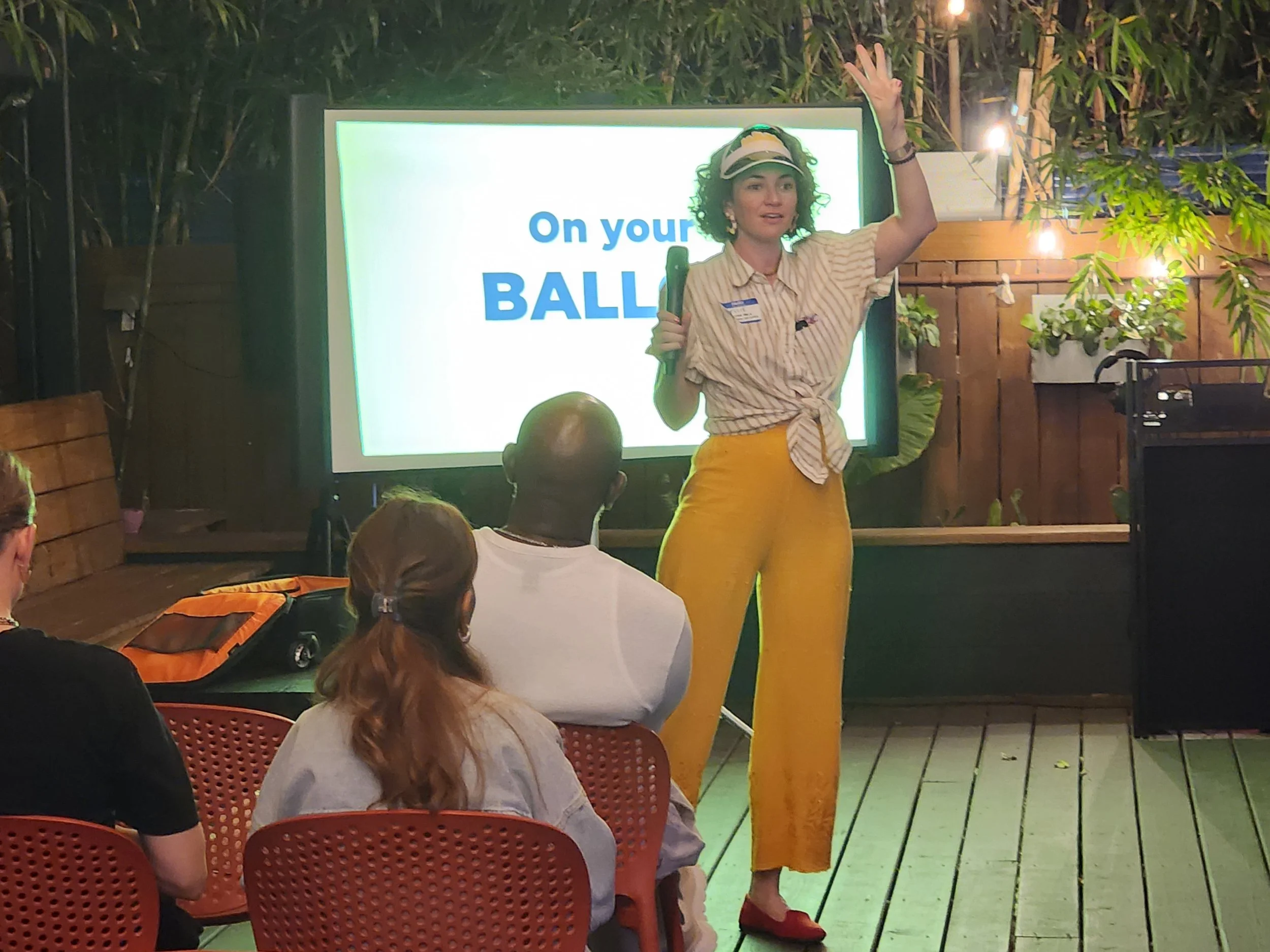 A woman with curly hair wearing a striped shirt, yellow pants, and a visor stands in front of a screen that says "On your BALI" while speaking to an audience seated in red chairs. The setting appears to be indoors with plants and warm lighting.