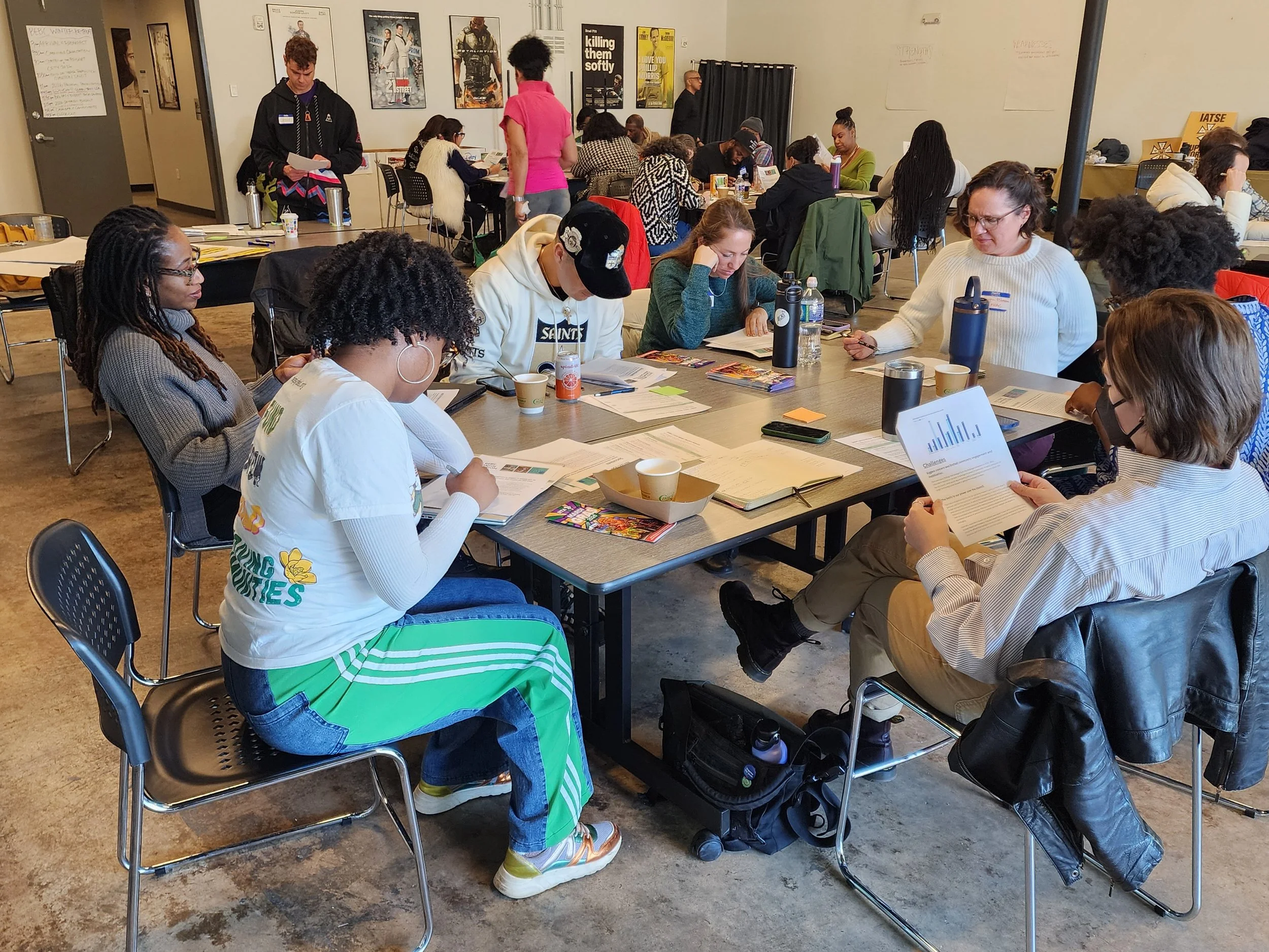 A diverse group of people sitting on chairs and tables in a room, engaged in reading, writing, and looking at documents, with some standing and walking around, posters on the wall, and various beverages on the tables.