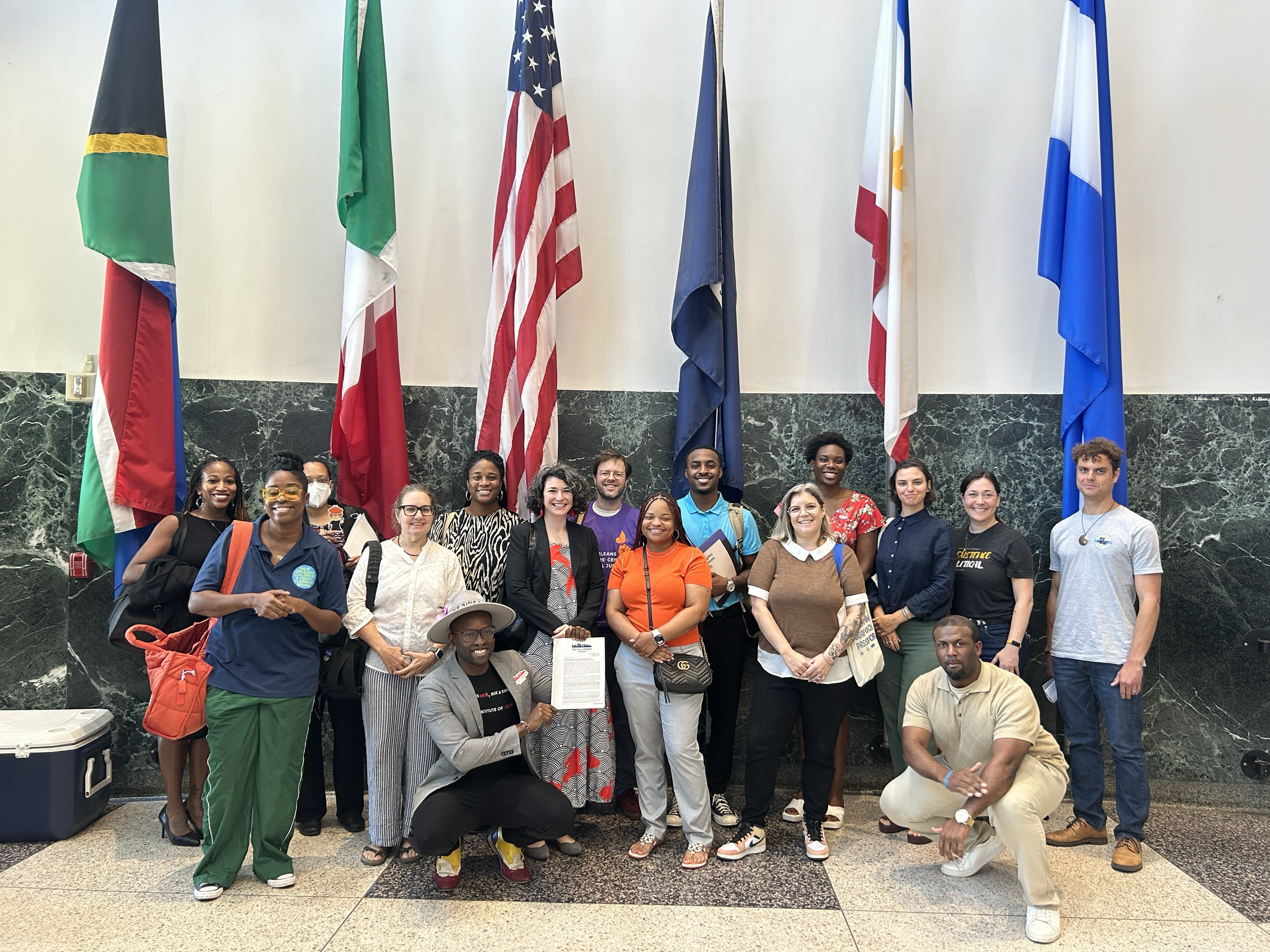 A diverse group of people gathered together indoors, smiling for a photo, with flags behind them representing different countries.