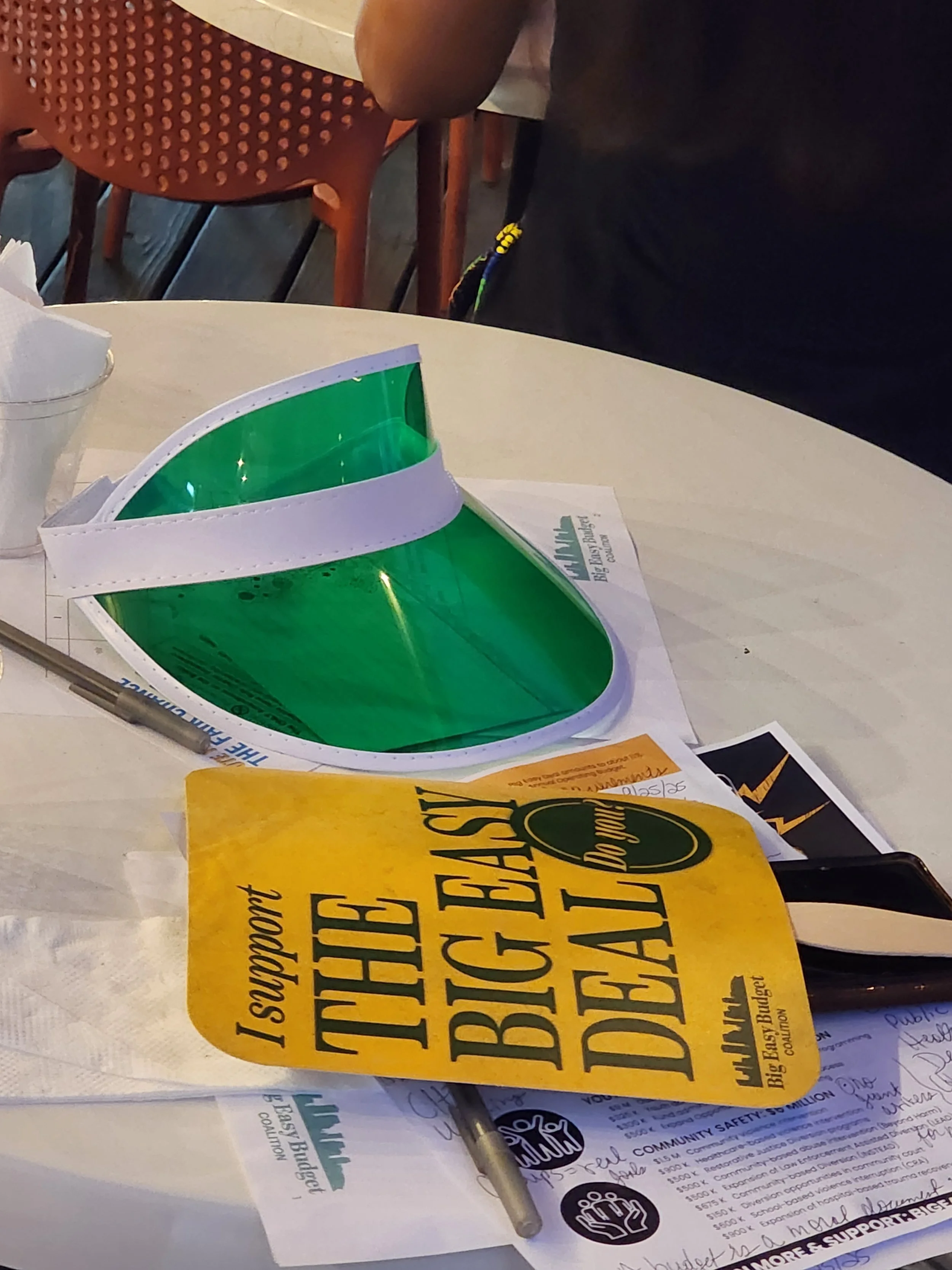A table with promotional flyers about community support, a yellow campaign flyer, a green and white sign, a pen, and some handwritten notes.