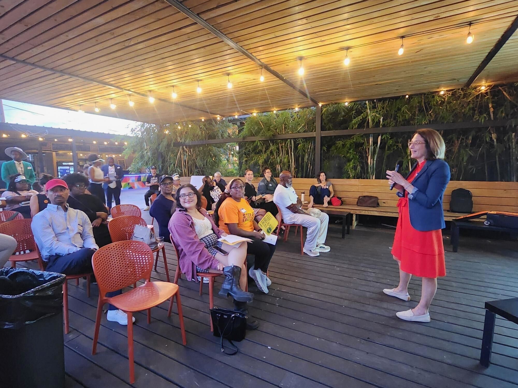 A woman in a blue blazer and red dress speaking into a microphone during an outdoor event, with seated audience members on wooden and red plastic chairs beneath a wooden ceiling with string lights, and green plants in the background.