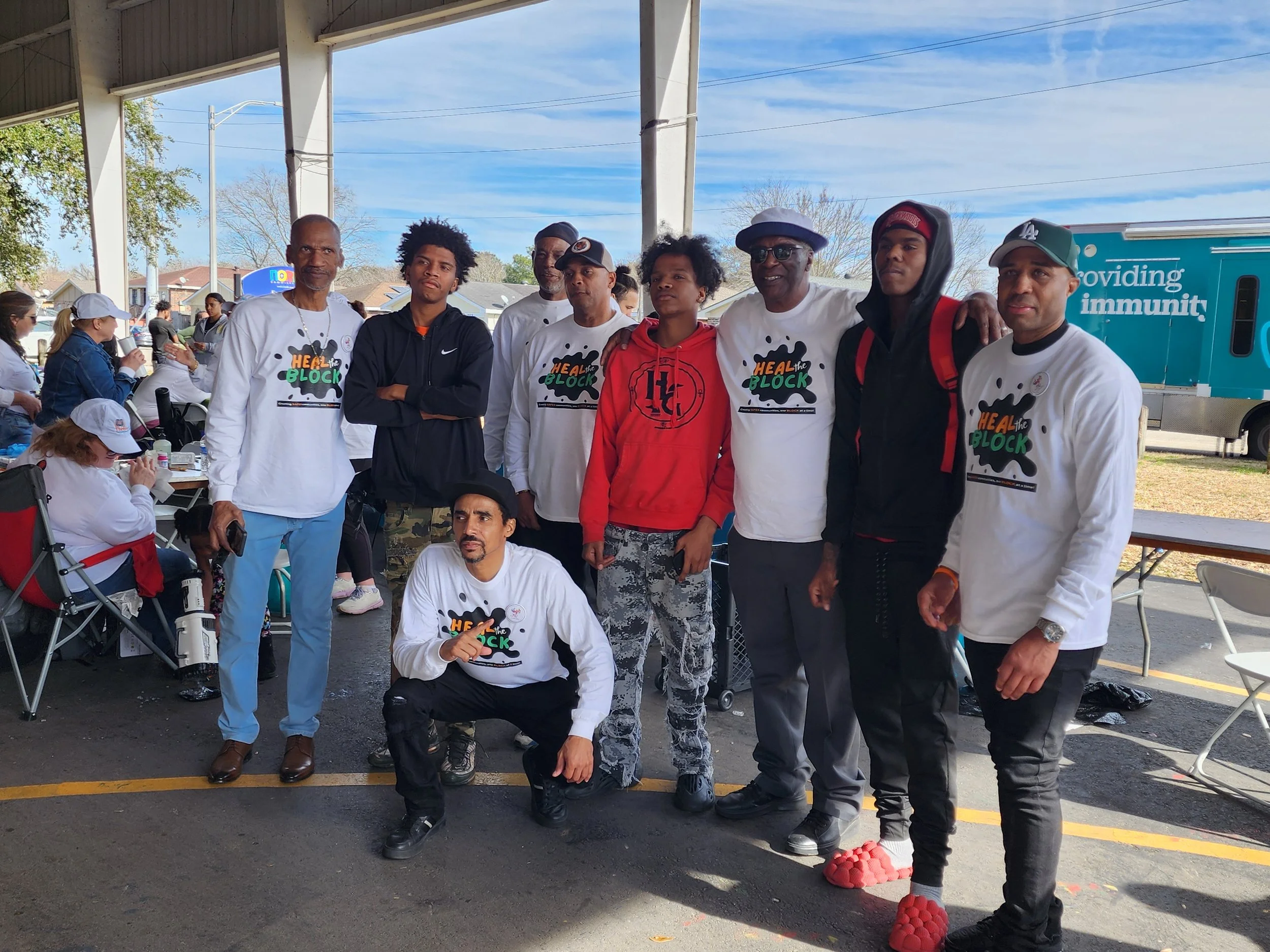 A group of nine people standing outdoors under a shelter, some wearing white shirts with 'Heal the Block' printed on them, with a food truck and people in the background.