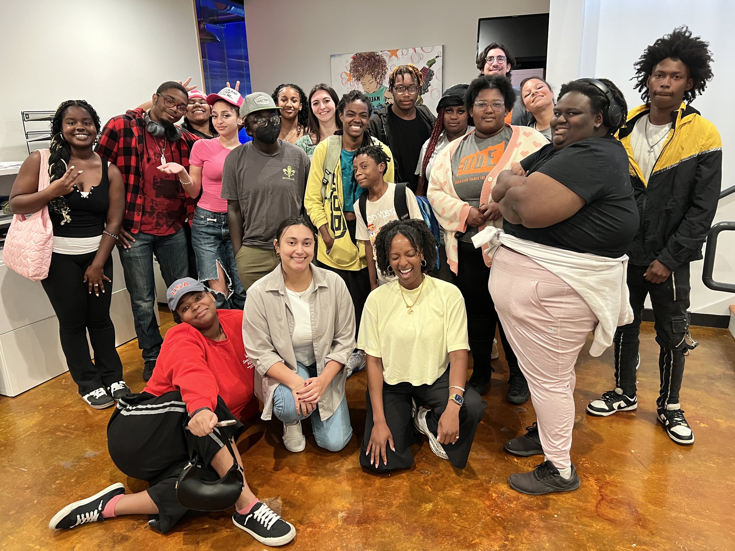 A diverse group of people posing inside a room with a wooden floor, some smiling, some making peace signs, and wearing casual clothing and accessories.