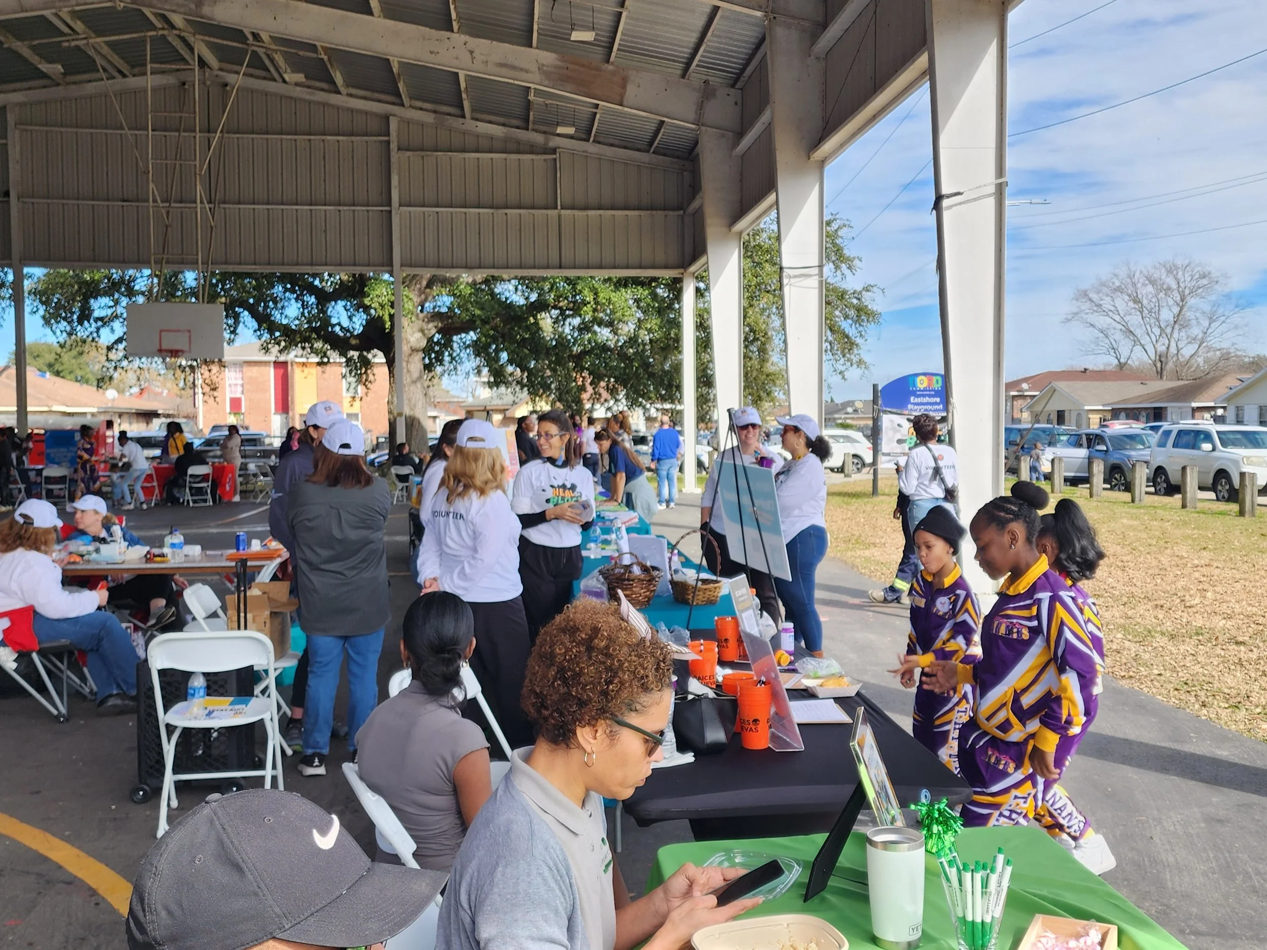 An outdoor community event with people gathered under a covered area. There are tables with items and volunteers wearing shirts with event logos. Spectators and participants are interacting, and children are present. The background shows a parking lot, trees, and a blue sign.