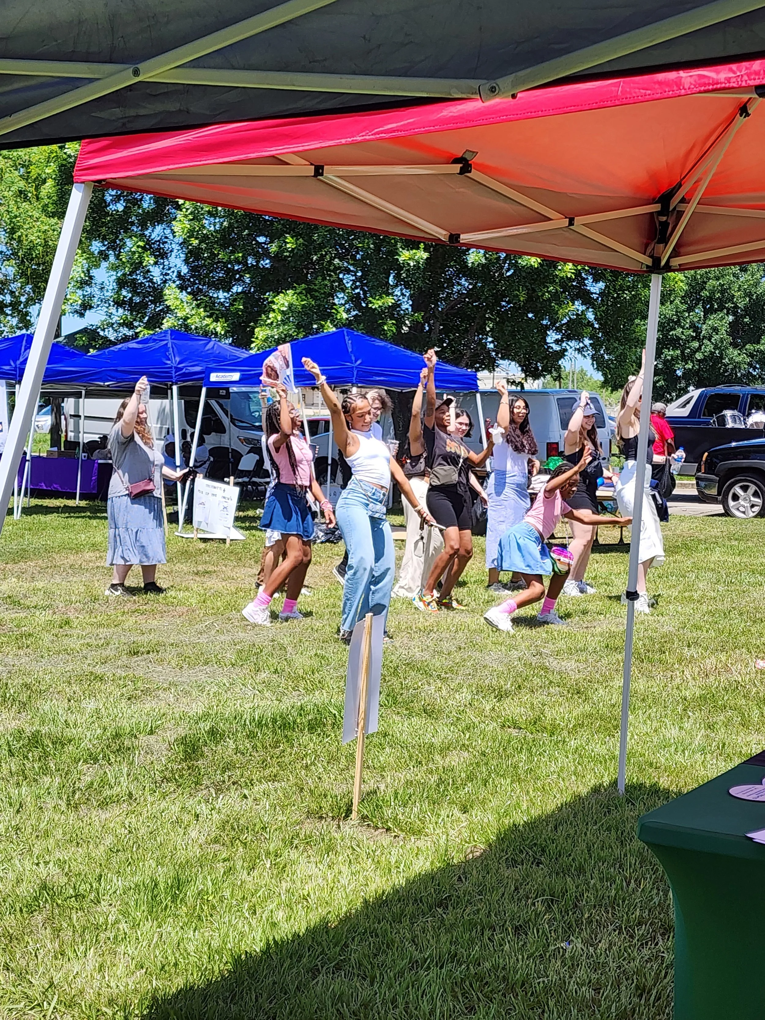 People standing on grass under tents, participating in an outdoor event on a sunny day, with trees and vehicles in the background.