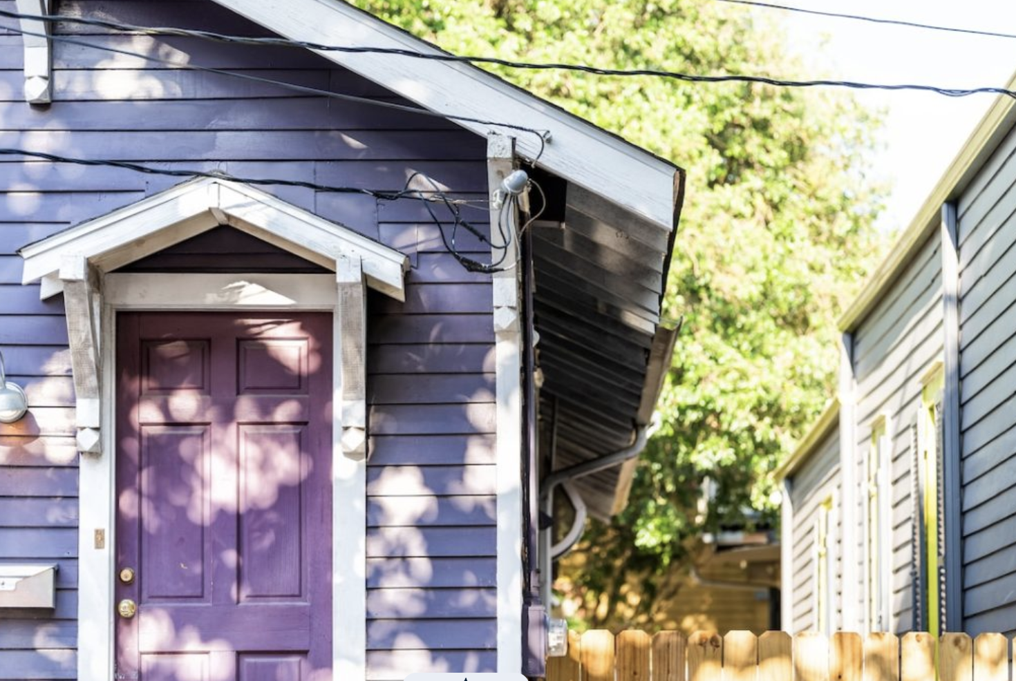 Close-up of a purple house with a purple door, white trim, and a small gable above the door. There are electrical wires and a light fixture visible on the house. Part of a neighboring house with blue siding and greenery in the background.