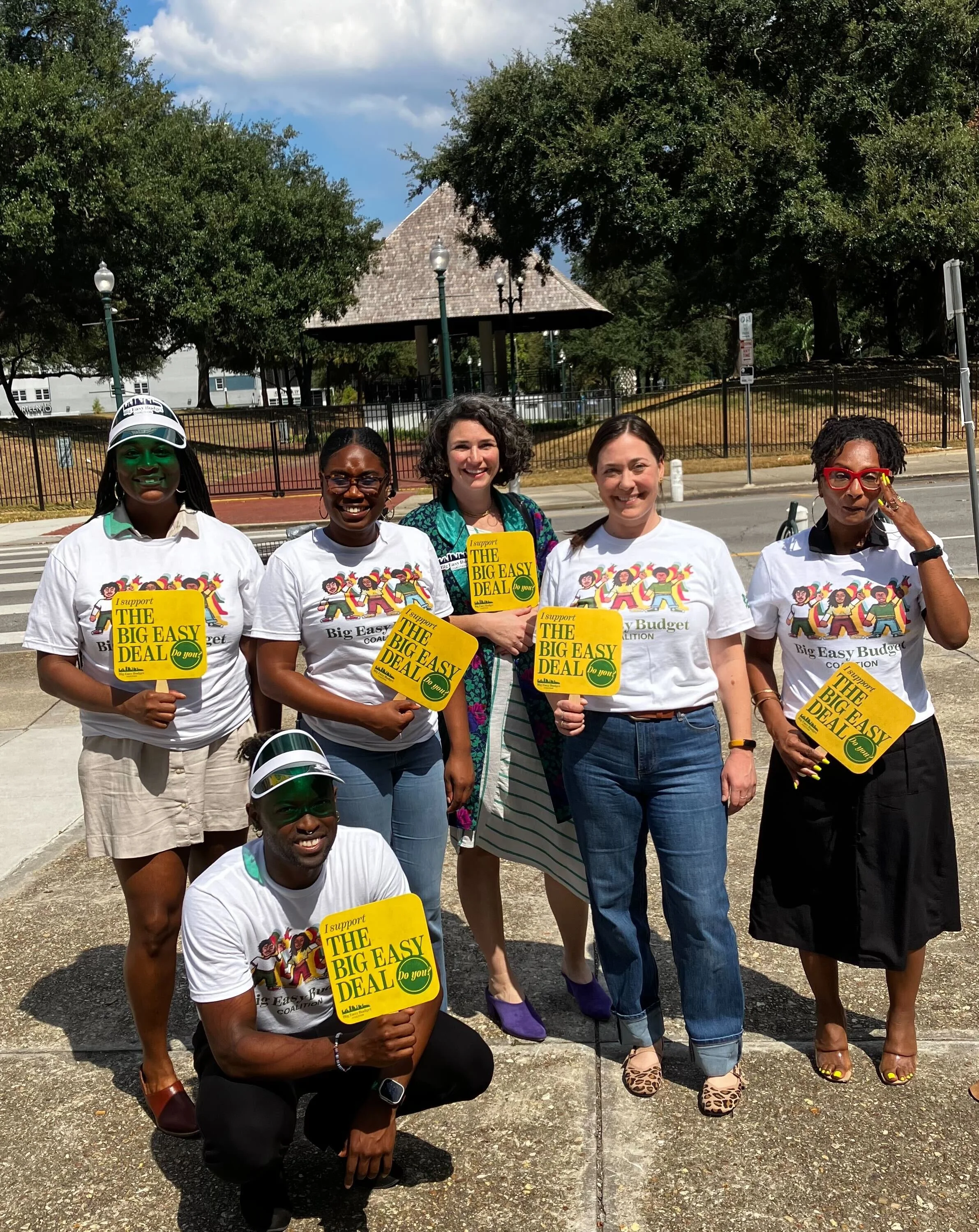 A group of people, some holding signs that say "I support THE BIG EASY DEAL do you?" standing outdoors on a sidewalk with trees and a gazebo in the background, during a sunny day.