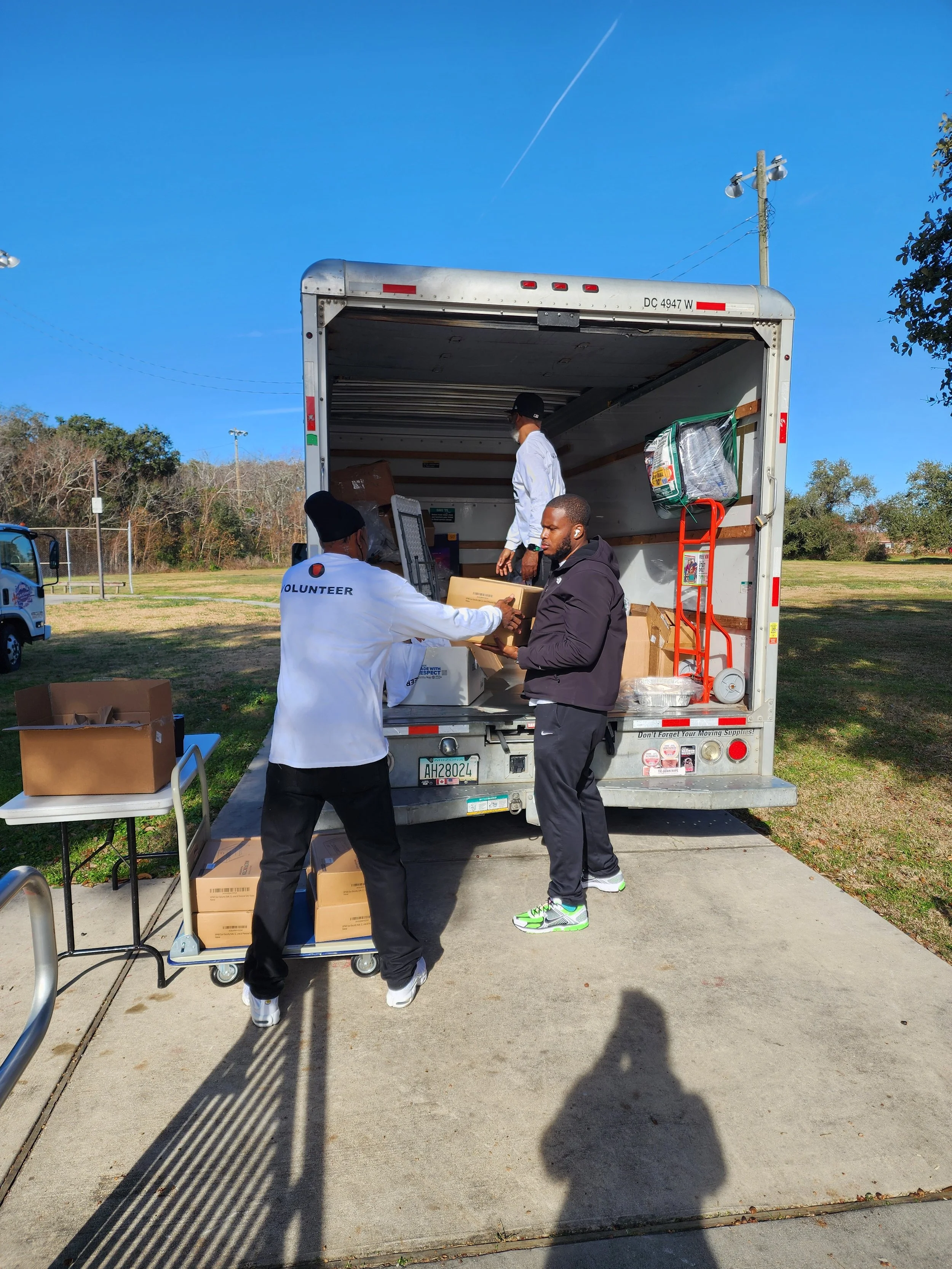 Three men are loading boxes into a truck at an outdoor park with a clear blue sky. One man is wearing a white jacket labeled 'Volunteer,' another in dark clothing, and the third in a white shirt and black hat.