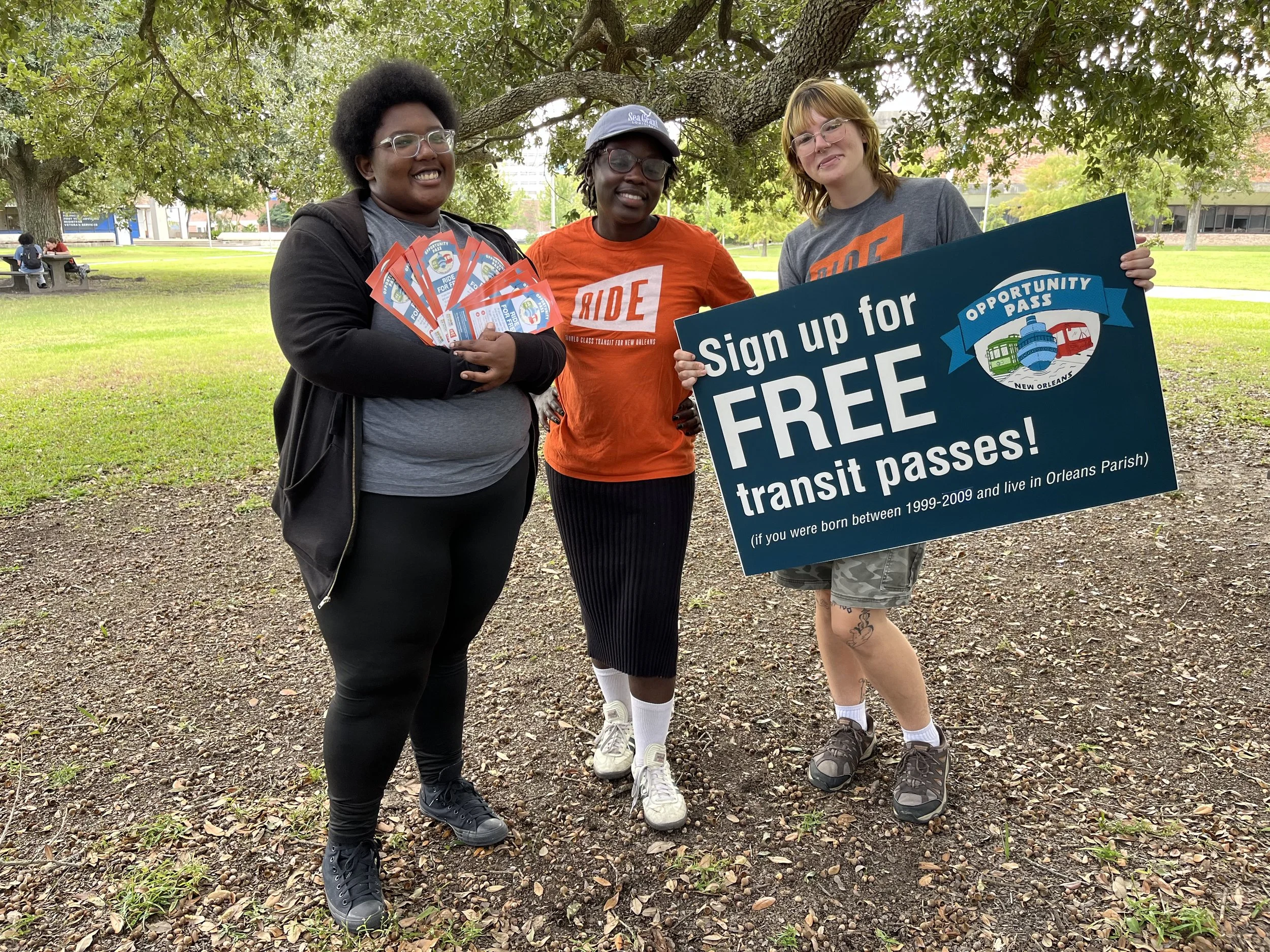 Three diverse people standing under a tree in a park, holding flyers and a large sign that promotes free transit passes in New Orleans.