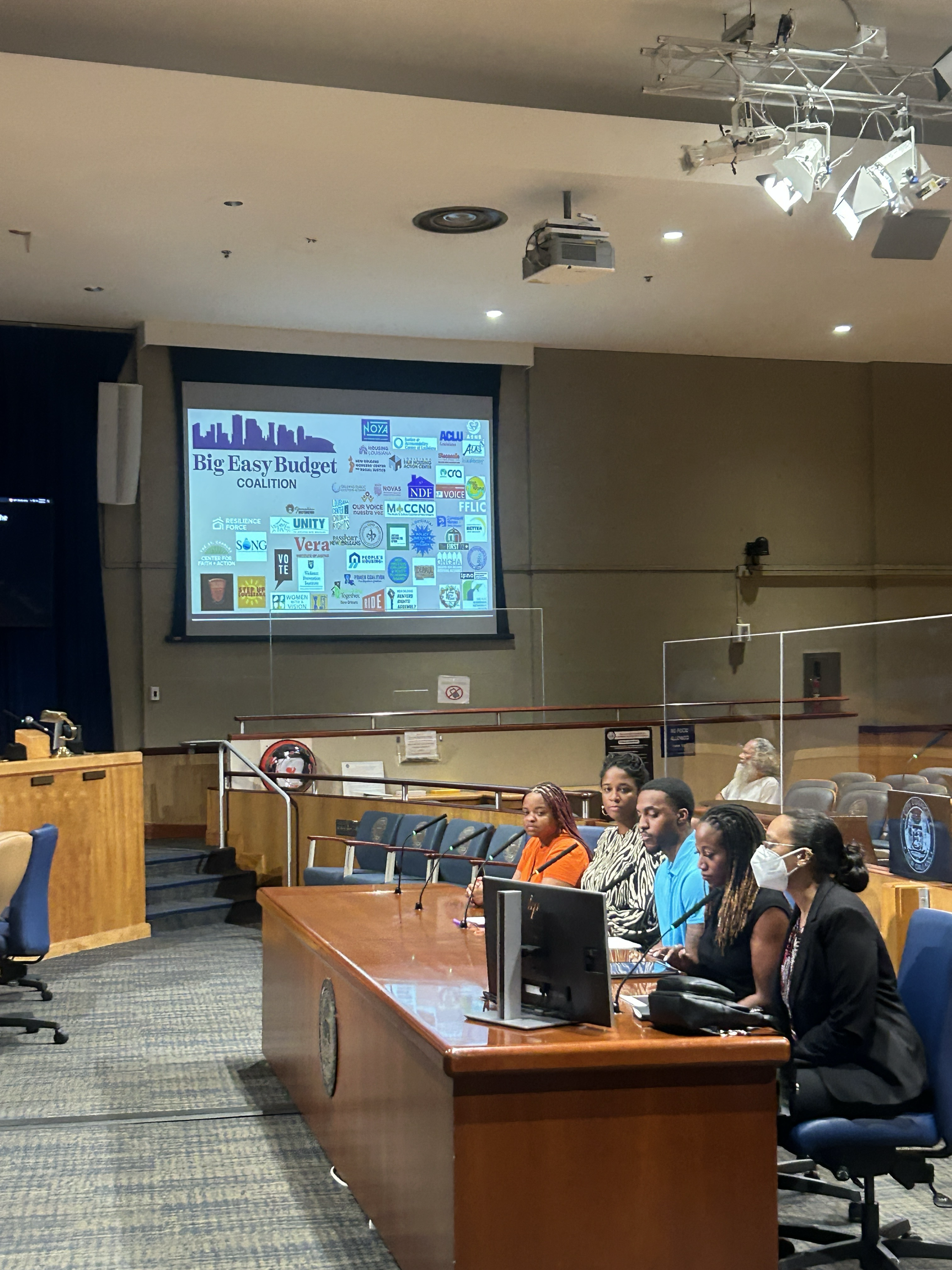 A group of people sitting at a long wooden conference table in a room with a large screen displaying a presentation titled "Big Easy Budget Coalition." The room appears to be a conference or meeting room with ceiling lights, a projection system, and a few individuals wearing face masks.