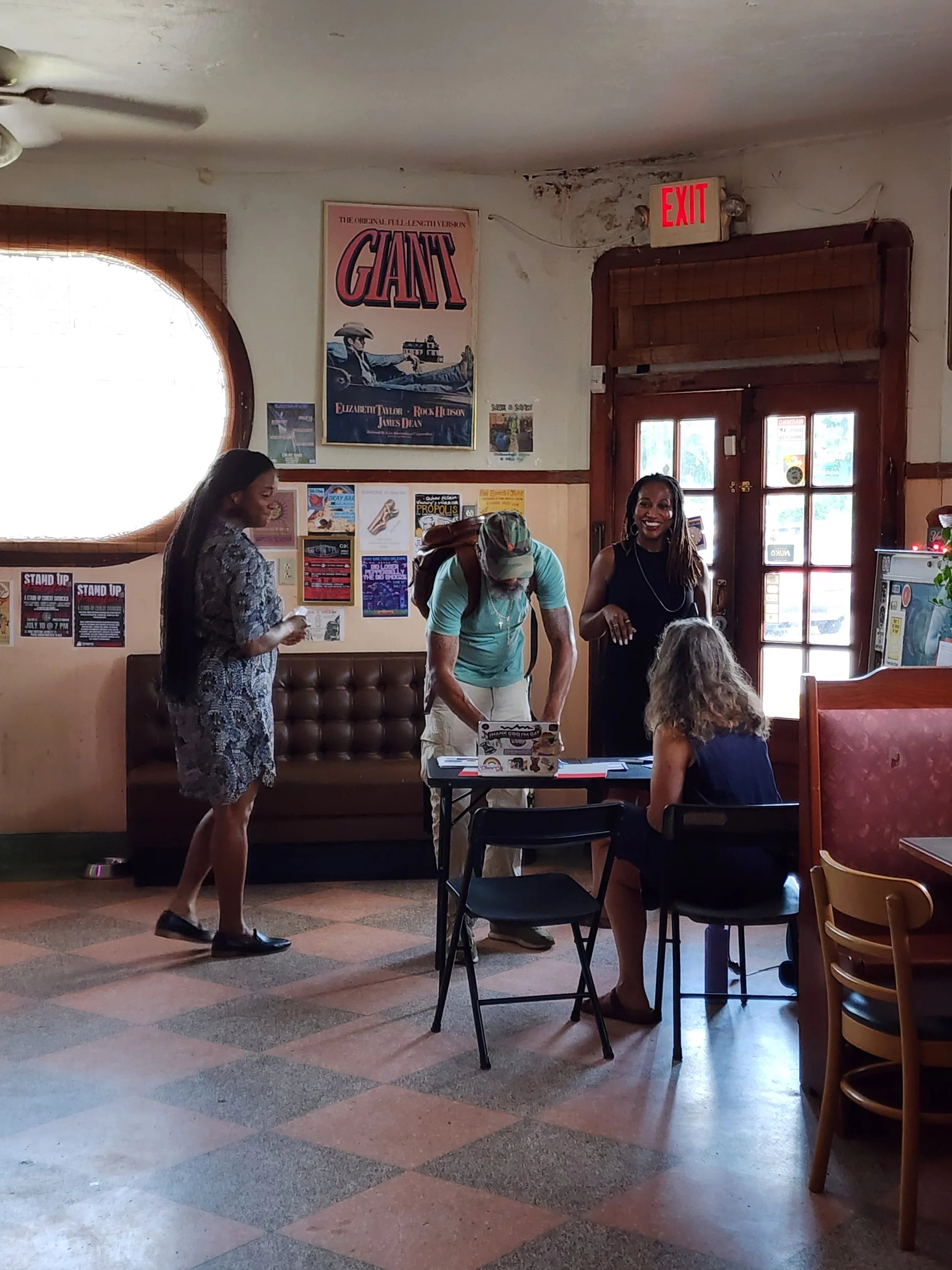 A group of people inside a cozy restaurant or cafe, with posters on the wall, including a vintage movie poster for 'Giant.' Three women and one man are interacting, with one woman standing, another woman sitting, a man leaning over a table, and the fourth woman smiling. The interior has wooden accents, a window with a rounded top, and an entrance door with glass panes. The floor is checkered with brown, orange, and gray tiles.