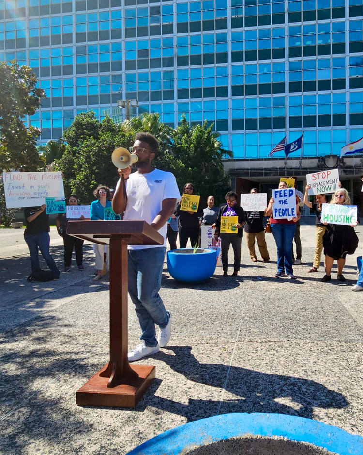A man speaking into a megaphone at a protest with people holding signs behind him in an outdoor urban setting, with a tall building and flags in the background.