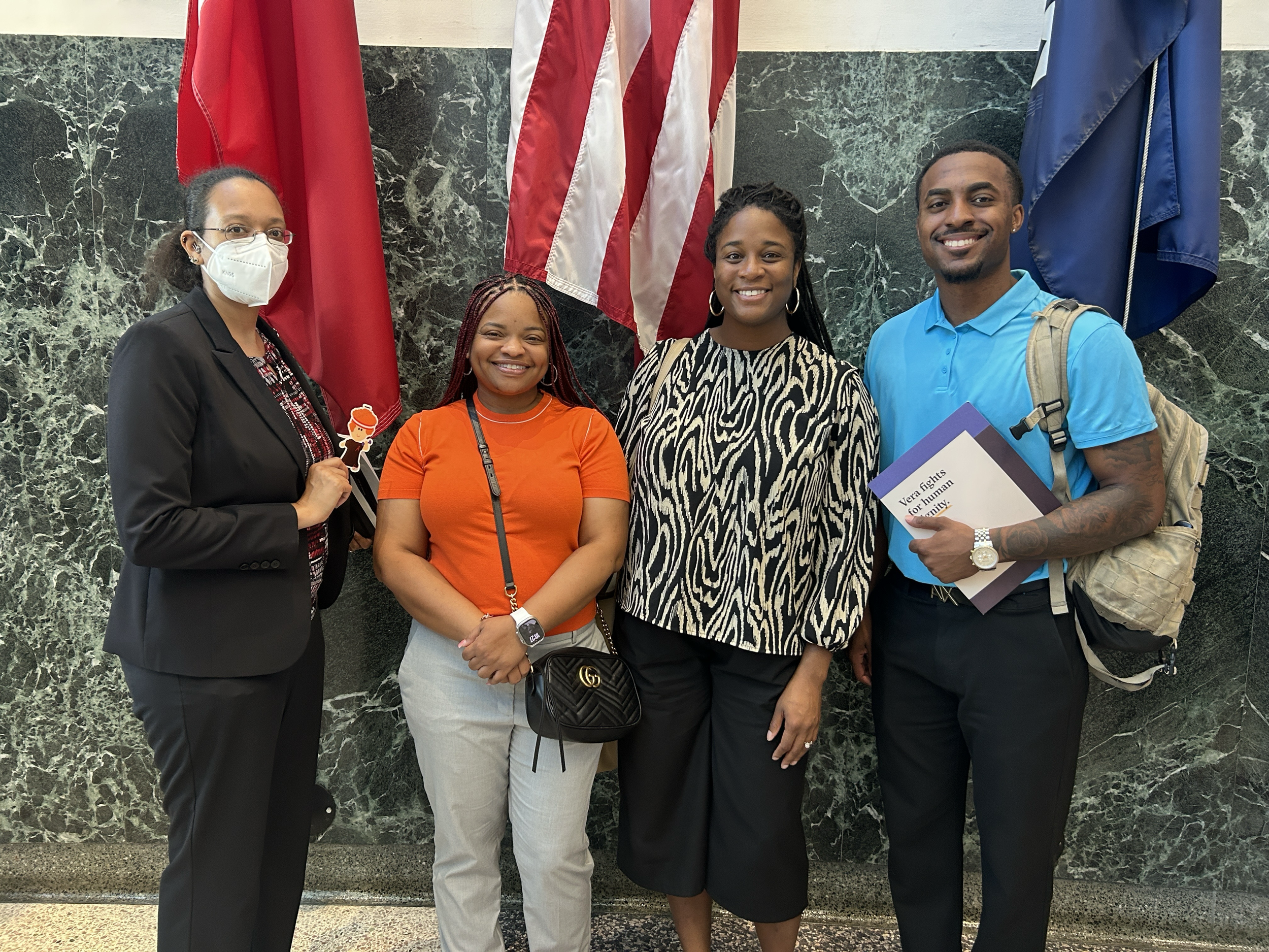 Group of four diverse people standing in front of three flags, smiling at the camera. One person is holding a small cartoon character sign, another has a box related to human rights.
