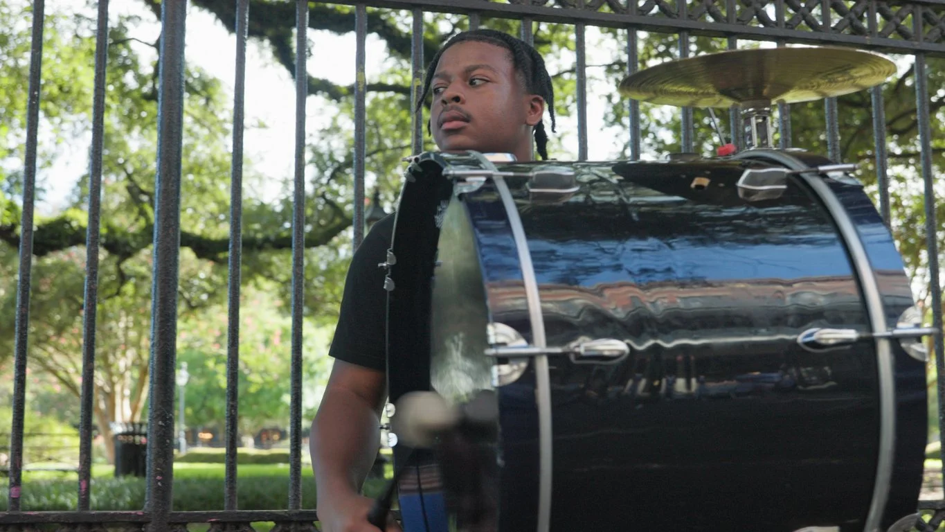 A young man with braided hair standing behind a black fence and looking to the side, with a large drum in front of him, outdoors in a park with trees.