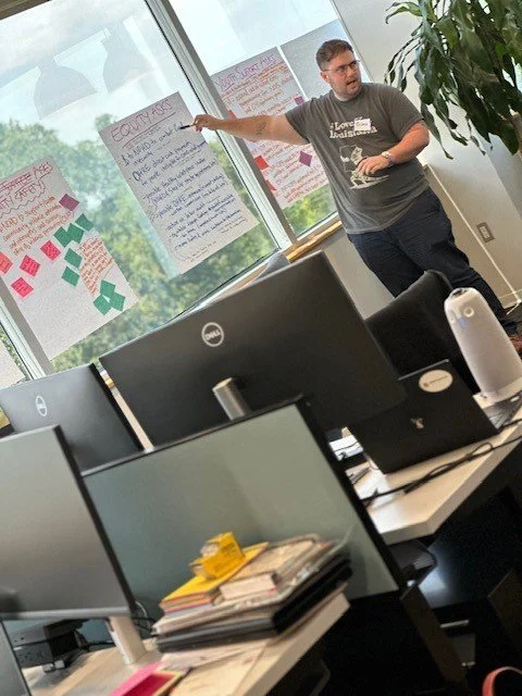 A man is standing near a window in an office, presenting a large paper with handwritten notes and post-it notes on it. The office has multiple computer monitors on desks, and there are multiple posters with notes and sticky notes on the window.