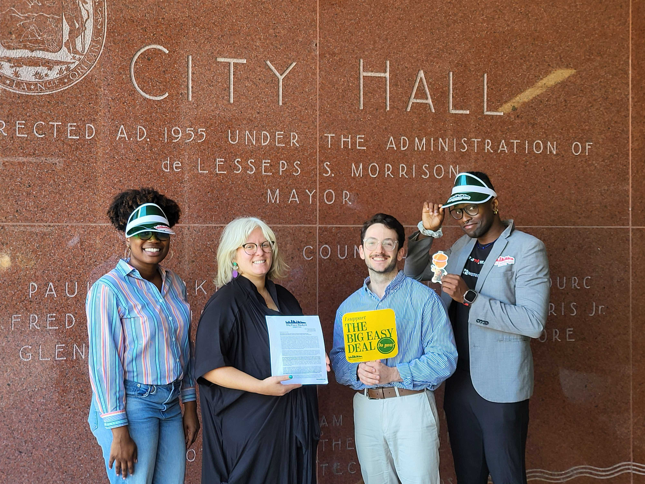 A group of five diverse people standing in front of a red marble wall with engraved text about the city hall. They are smiling, holding signs and papers, and some are wearing sunglasses and hats.