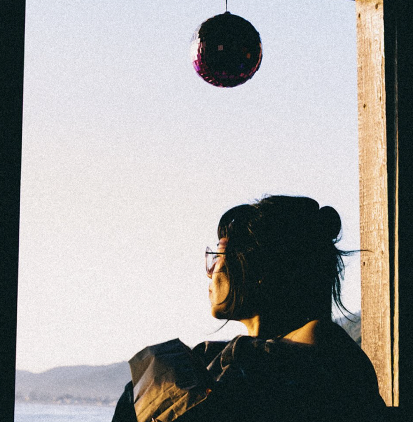 Phoebe Geonzon in profile, looking out a window of a Steep Ravine cabin toward the Pacific Ocean, with a sunlit disco ball hanging above. Photo by Dani Nomura.