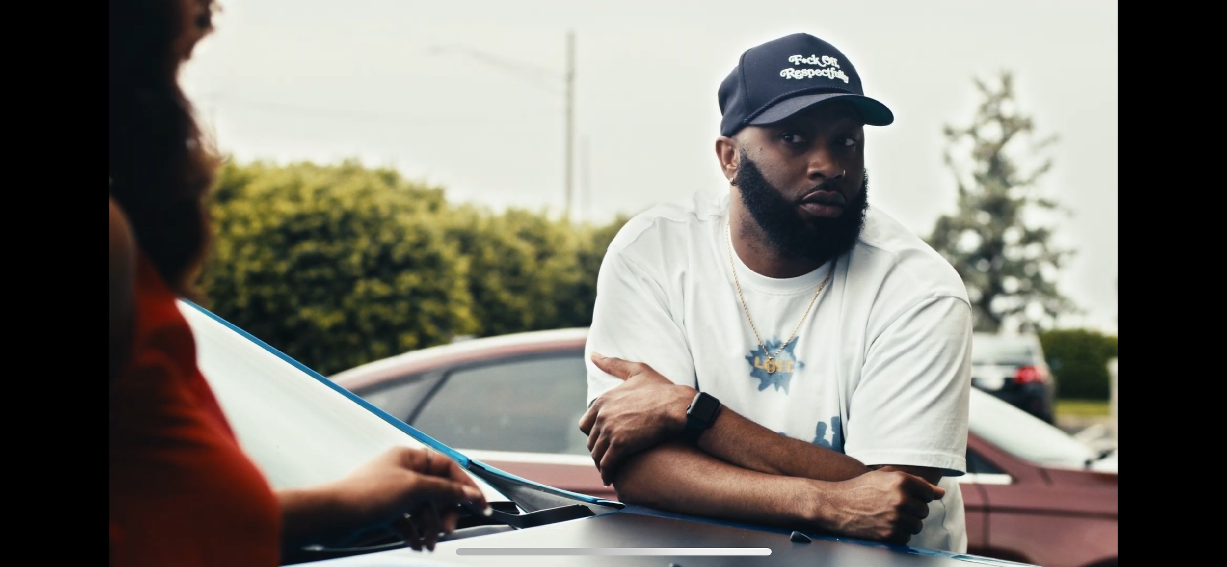 A man with a beard wearing a black cap that says 'Pack Oil Respectfully,' a white t-shirt, and a necklace with a 'LOL' pendant, is leaning on a car and talking to a person with curly hair in a red shirt, who is writing in a notebook outdoors.