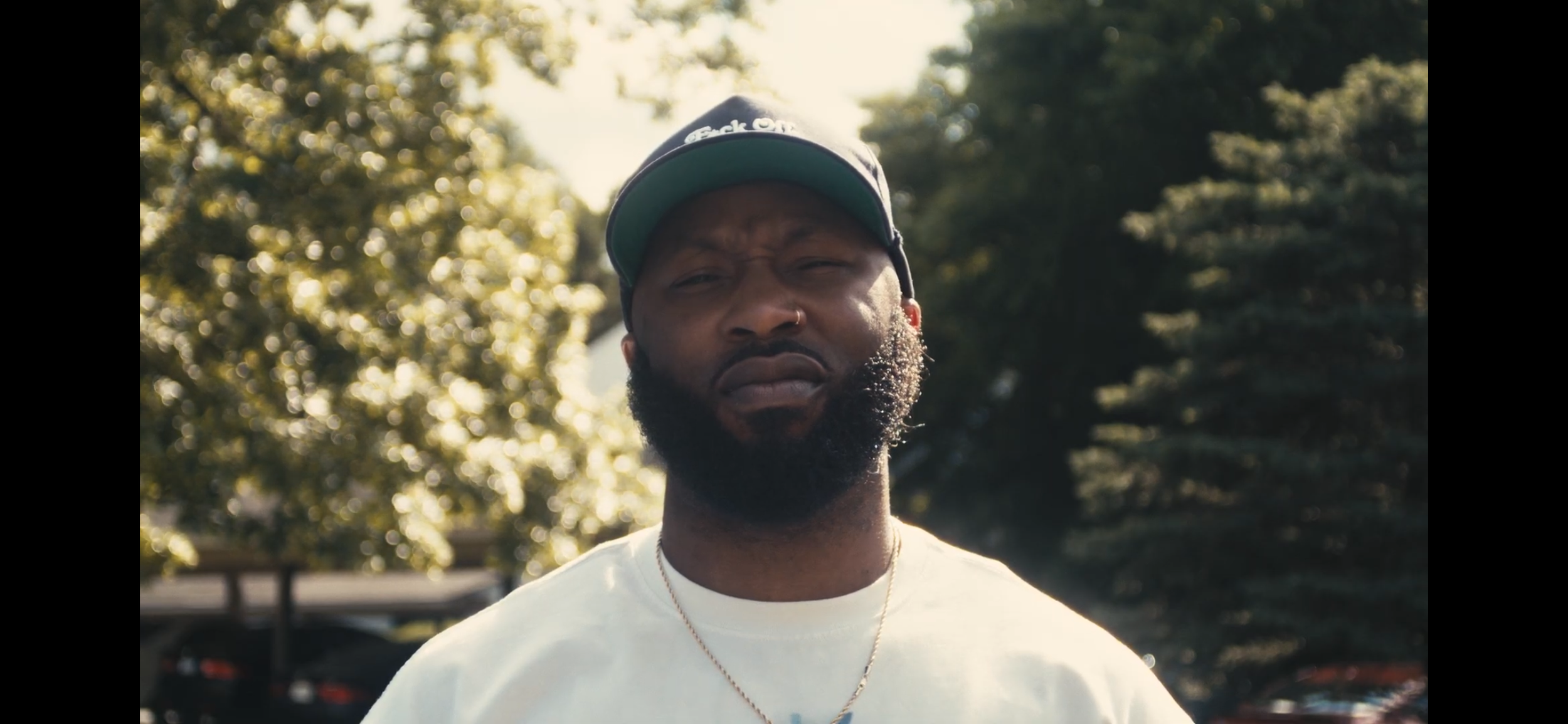 Close-up of a young man with a beard wearing a baseball cap and a white t-shirt, outdoors with trees and sunlight in the background.