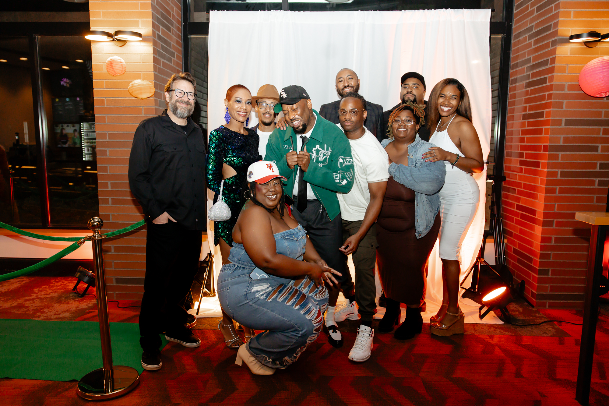 A diverse group of ten people, including men and women of different ages and ethnicities, smiling and posing together at an indoor social event. They are standing in front of a white backdrop with red brick walls on either side, some wearing casual and semi-formal clothing, and one person is kneeling in front.