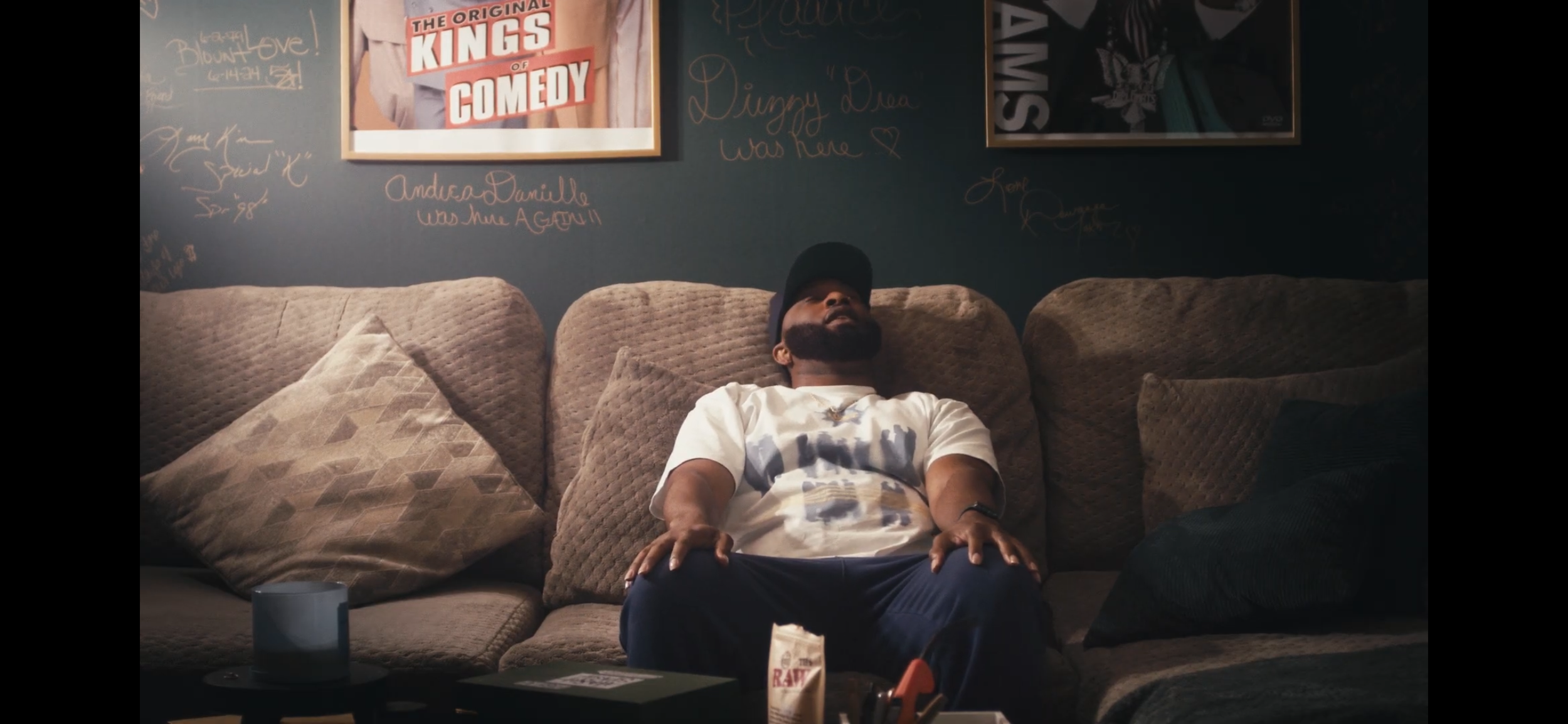 A man with a beard and wearing a cap, sitting on a beige couch with patterned pillows, appears to be sleeping or resting. The background features a dark wall with various signs and signatures, including a framed poster that says "The Original Kings o