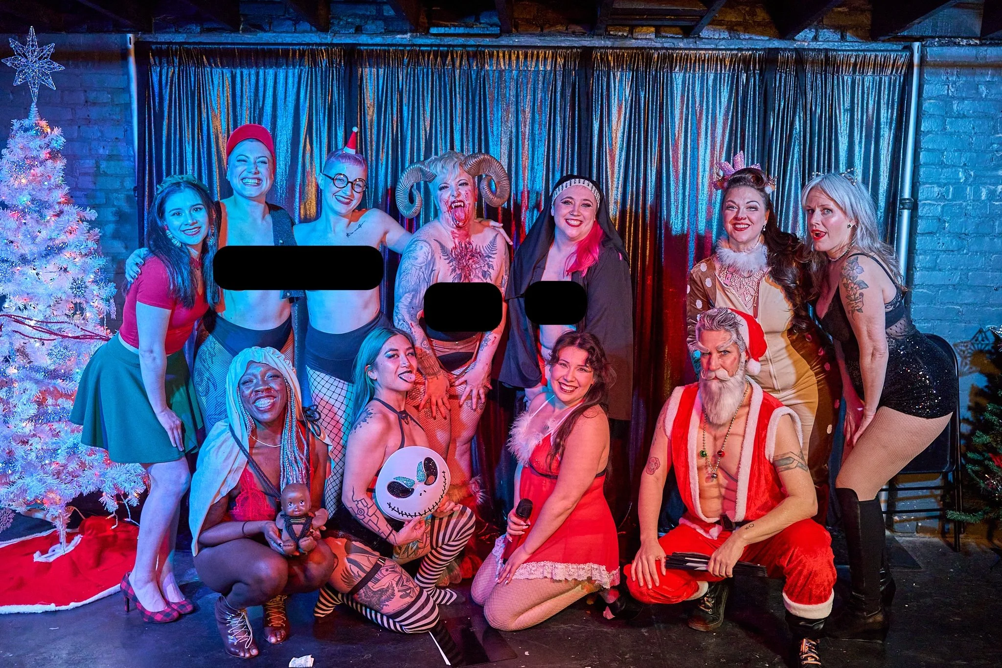 A group of people in festive burlesque costumes posing on a stage with a decorated Christmas tree, colorful lights, and a shiny curtain backdrop.