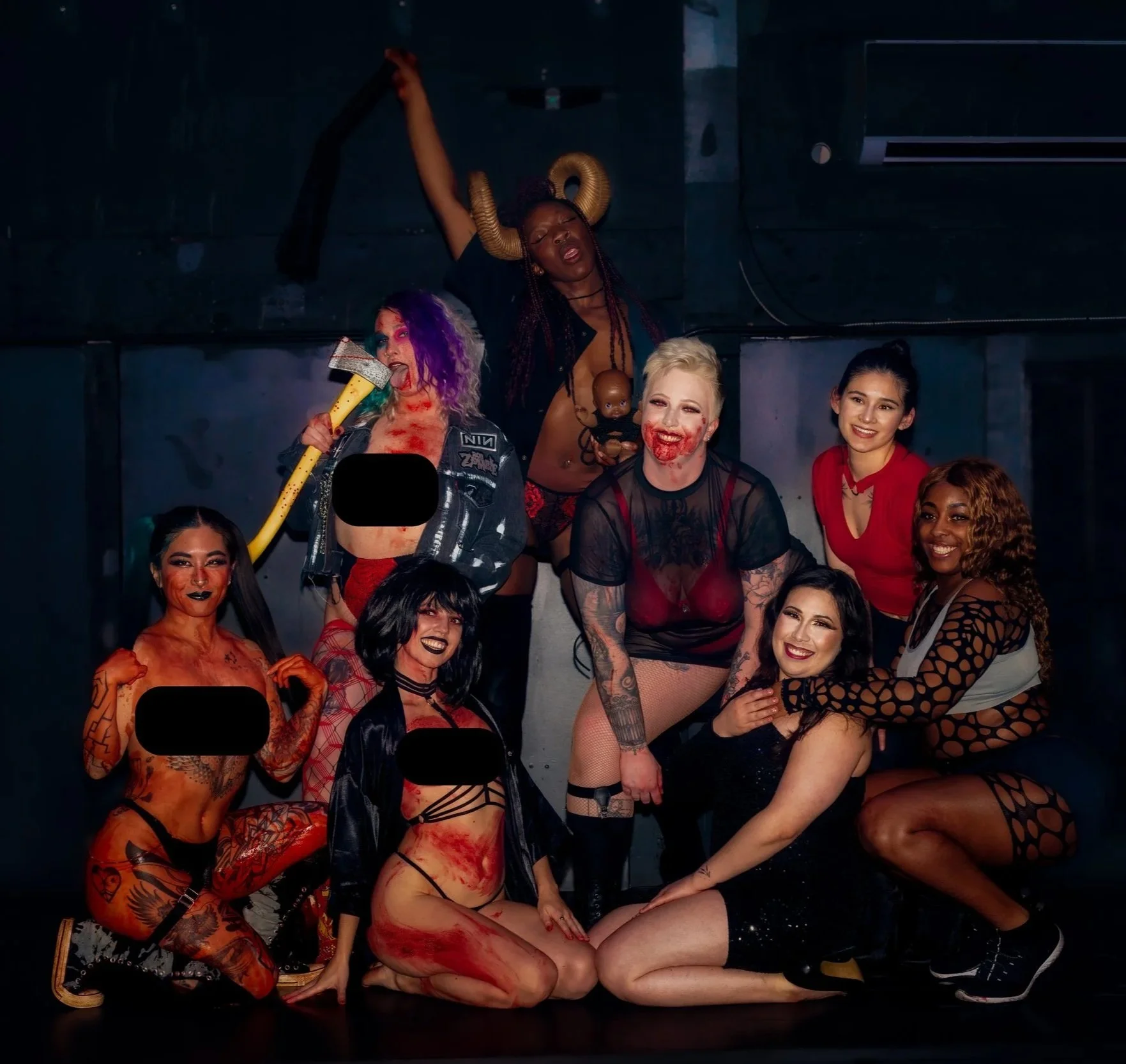 A diverse group of people dressed in provocative and Halloween-themed costumes posing together in a dark setting, some with makeup resembling blood and scars, with confident and playful expressions.