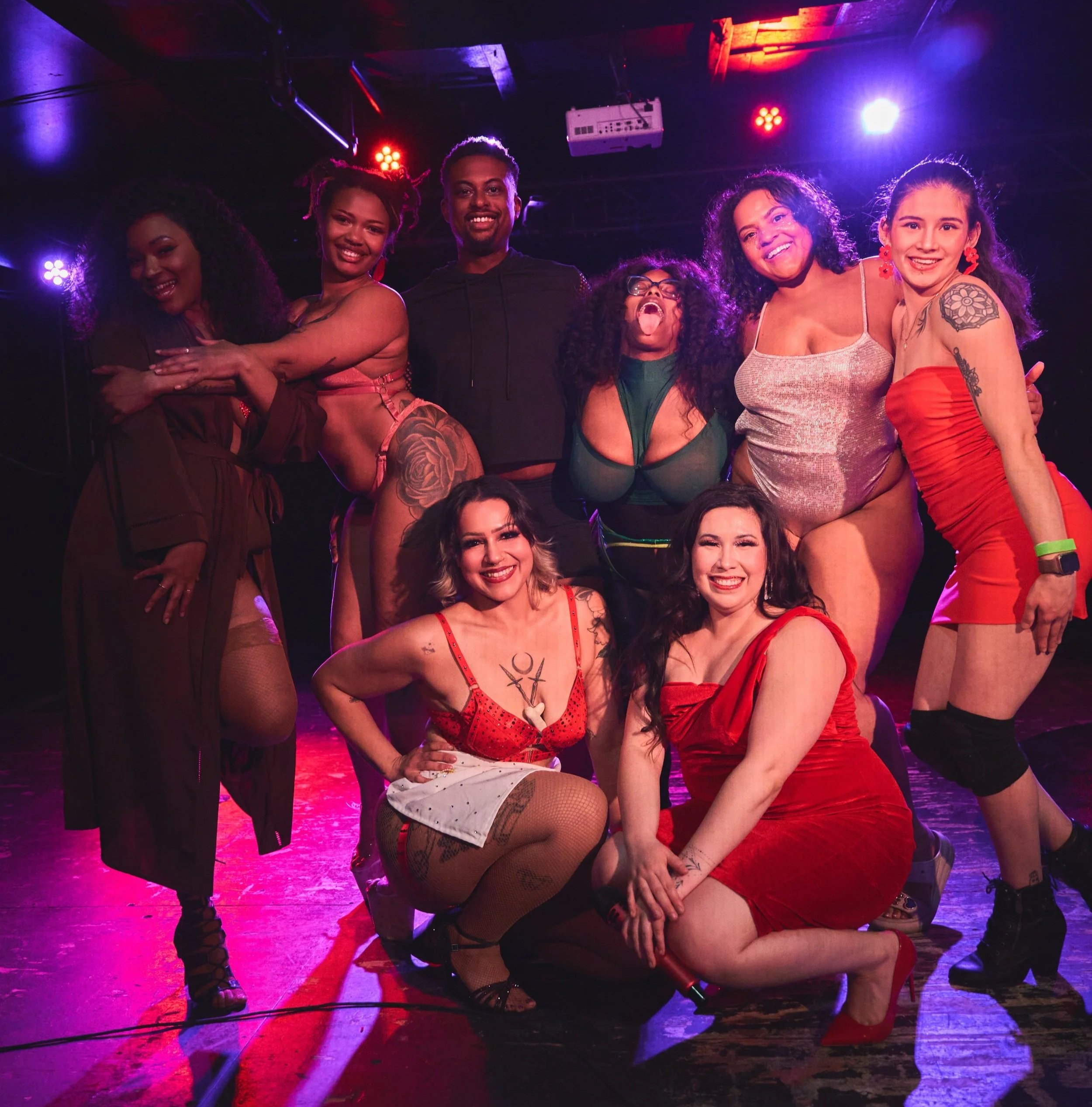 Group of eight people posing together on a stage with colorful stage lighting. They are dressed in burlesque outfits and showing tattoos