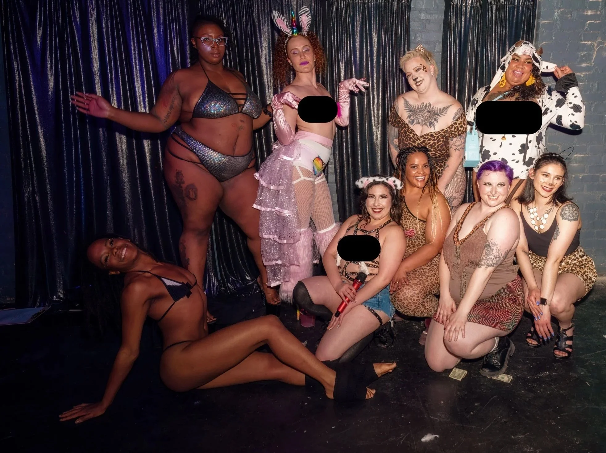 A group of nine diverse women posing together on stage with a black curtain background, some in animal-themed buroesque costumes and showing various expressions.