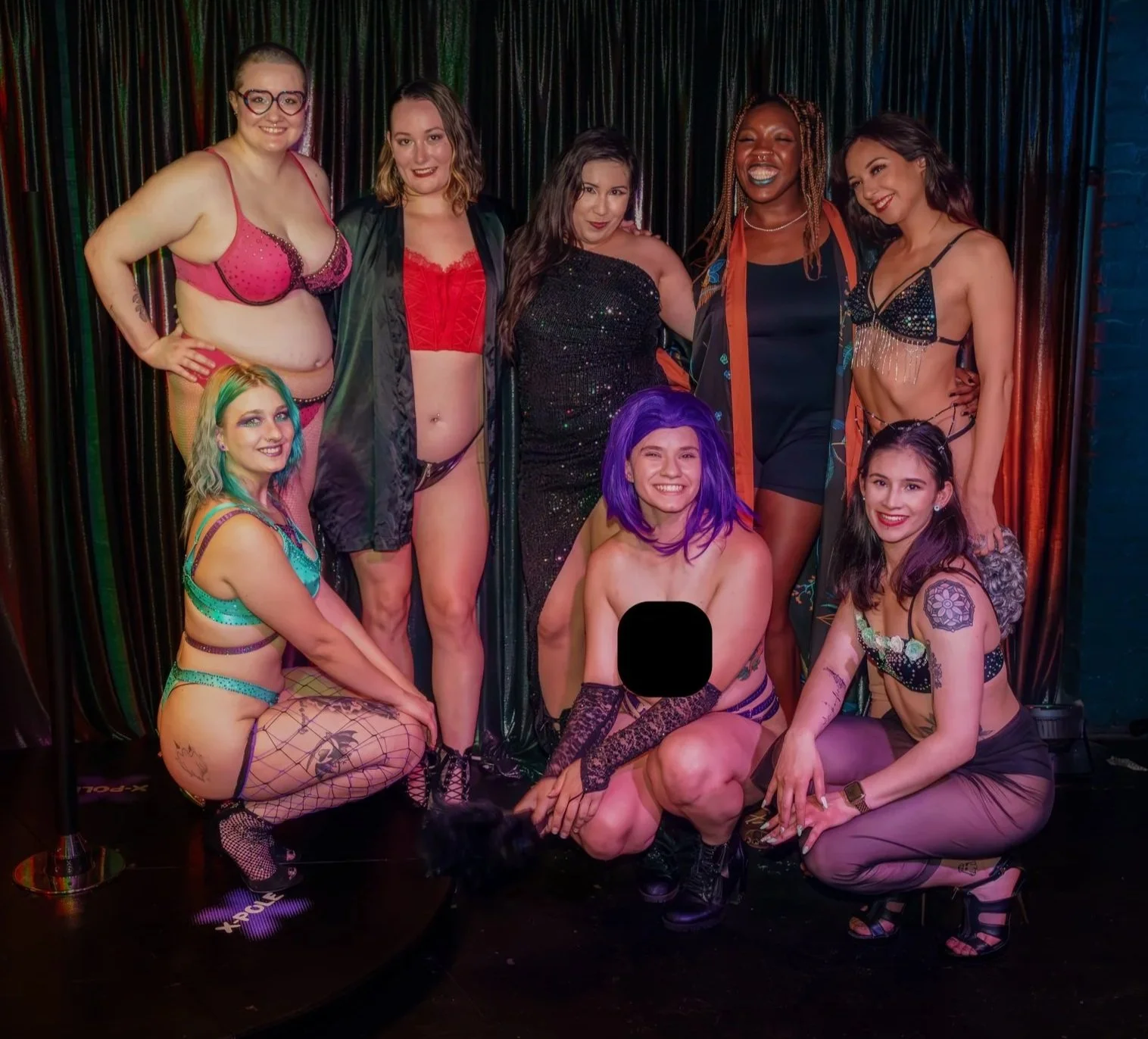 Group of women in colorful lingerie and costumes, smiling and posing on stage with a black curtain background at a festive event.