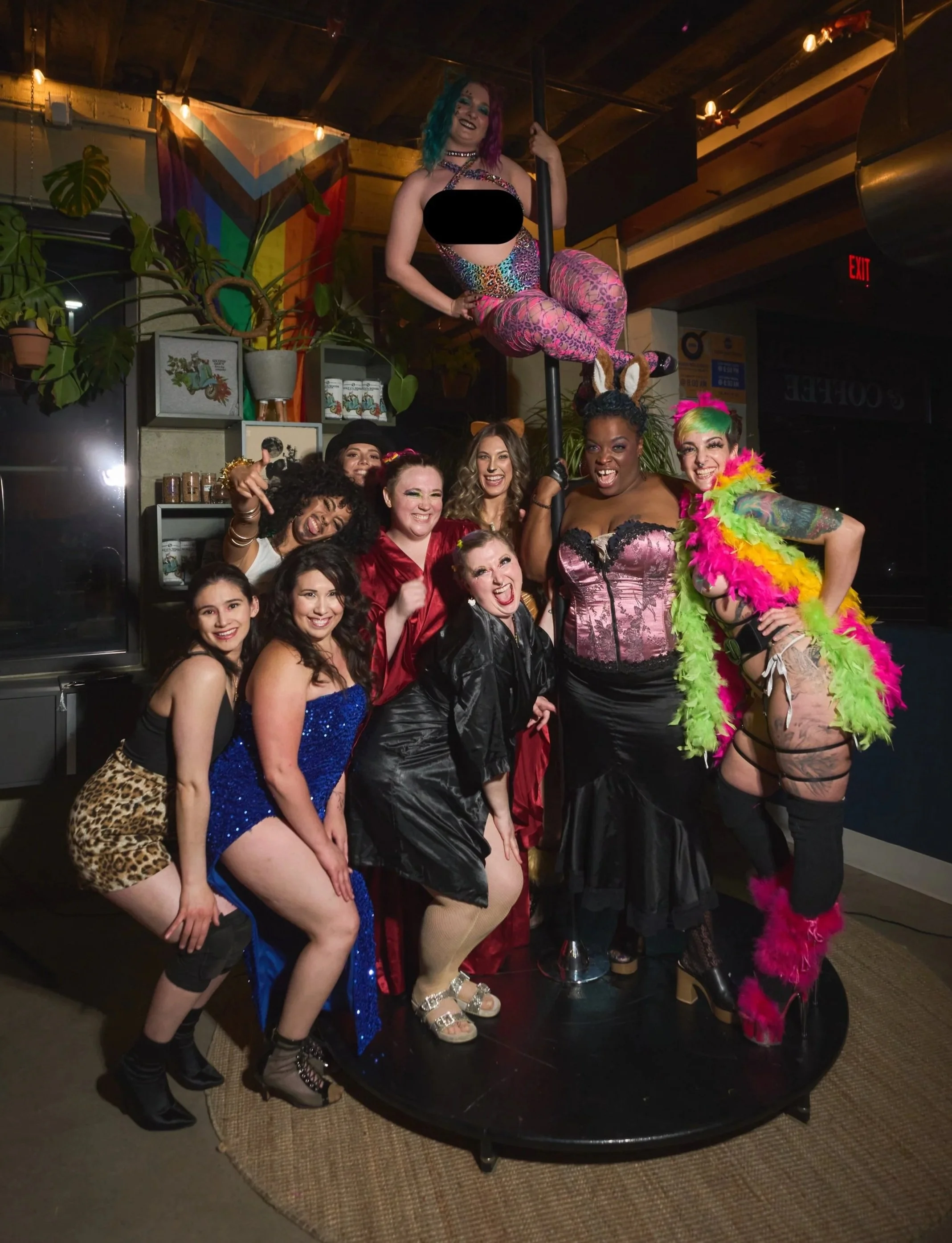 A group of women in colorful, shiny, and themed costumes posing around a pole in a lively indoor setting, with a woman on the pole dressed in rainbow-colored tights and a black top, while others wear various vibrant dresses and accessories, some with