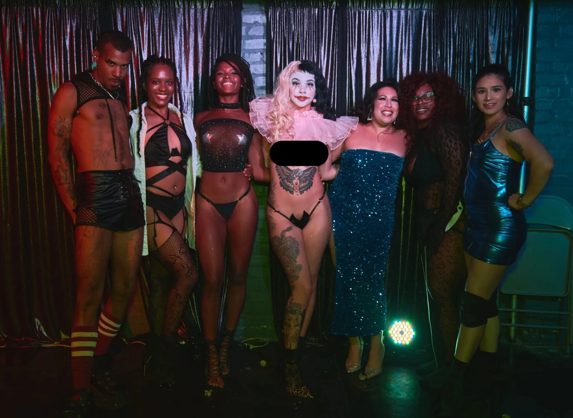 Group of diverse people in colorful and eclectic outfits posing together at a burlesque event, with a curtain backdrop and colorful lighting.
