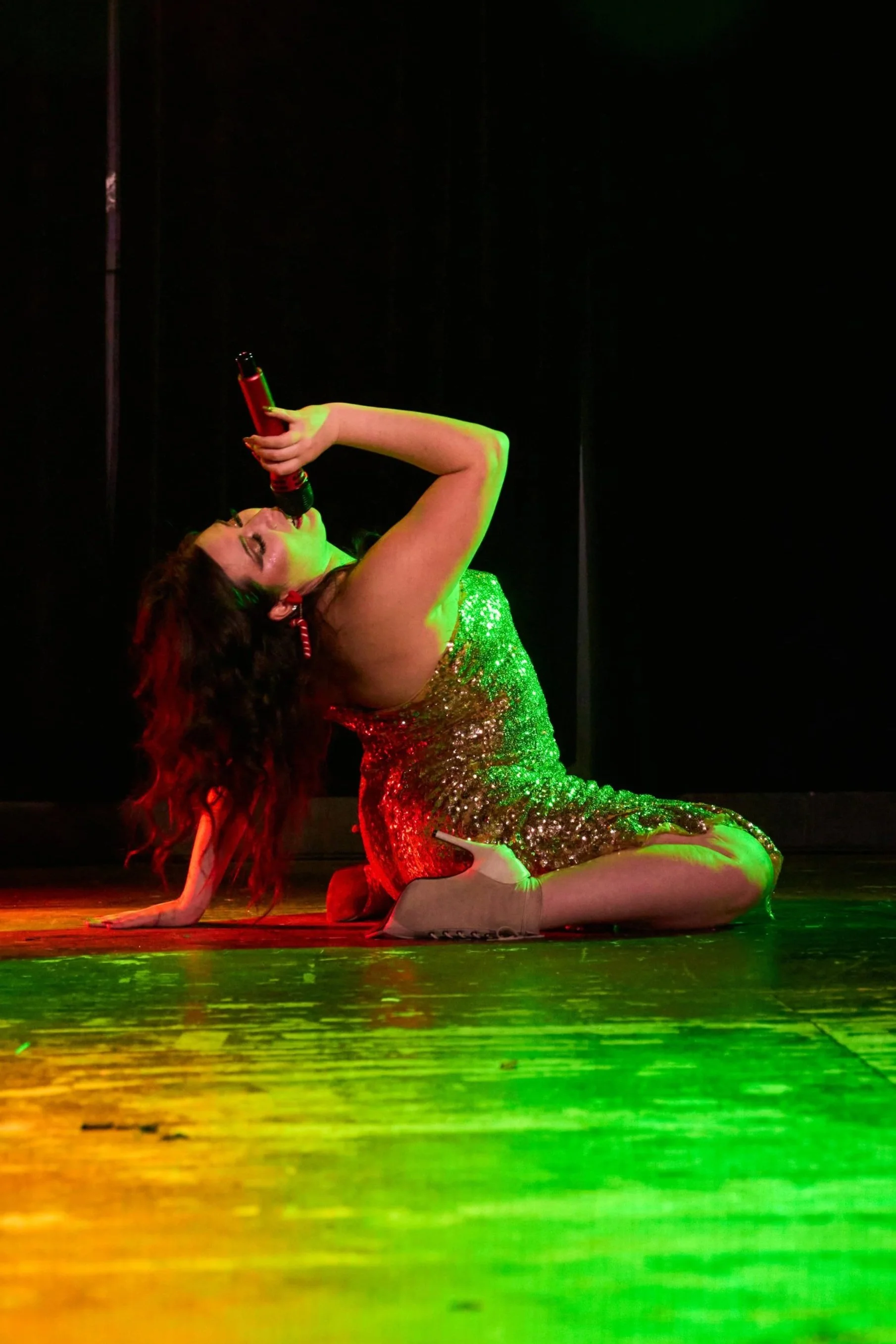 A female performer in a glittery dress is on stage, kneeling with one leg bent and the other extended behind her, holding a microphone and singing with eyes closed. The stage lighting casts a green and yellow glow.