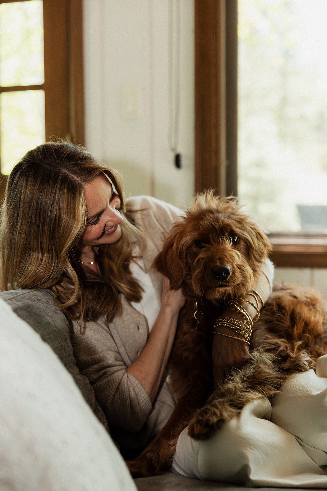 A woman with long wavy hair holding a cute brown dog with curly fur, sitting on a couch near a window.
