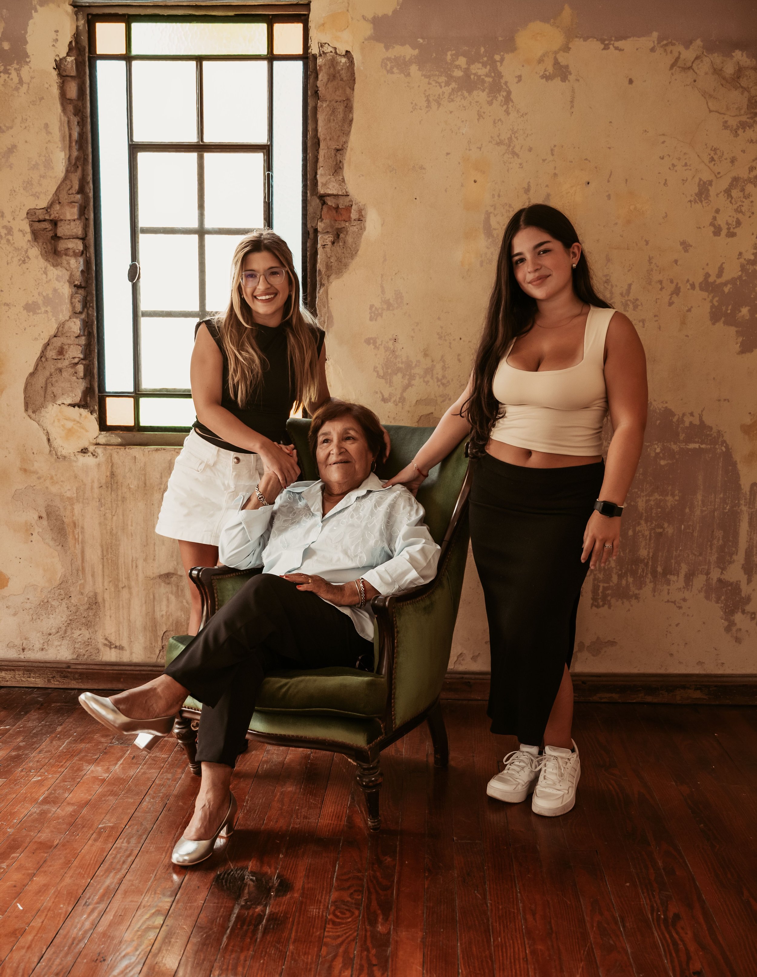 Three women, one elderly and two younger, posing together in a rustic room with peeling yellow paint and a wooden floor, near a window with colorful stained glass. The elderly woman is seated in a green armchair, wearing a white blouse, black pants, 