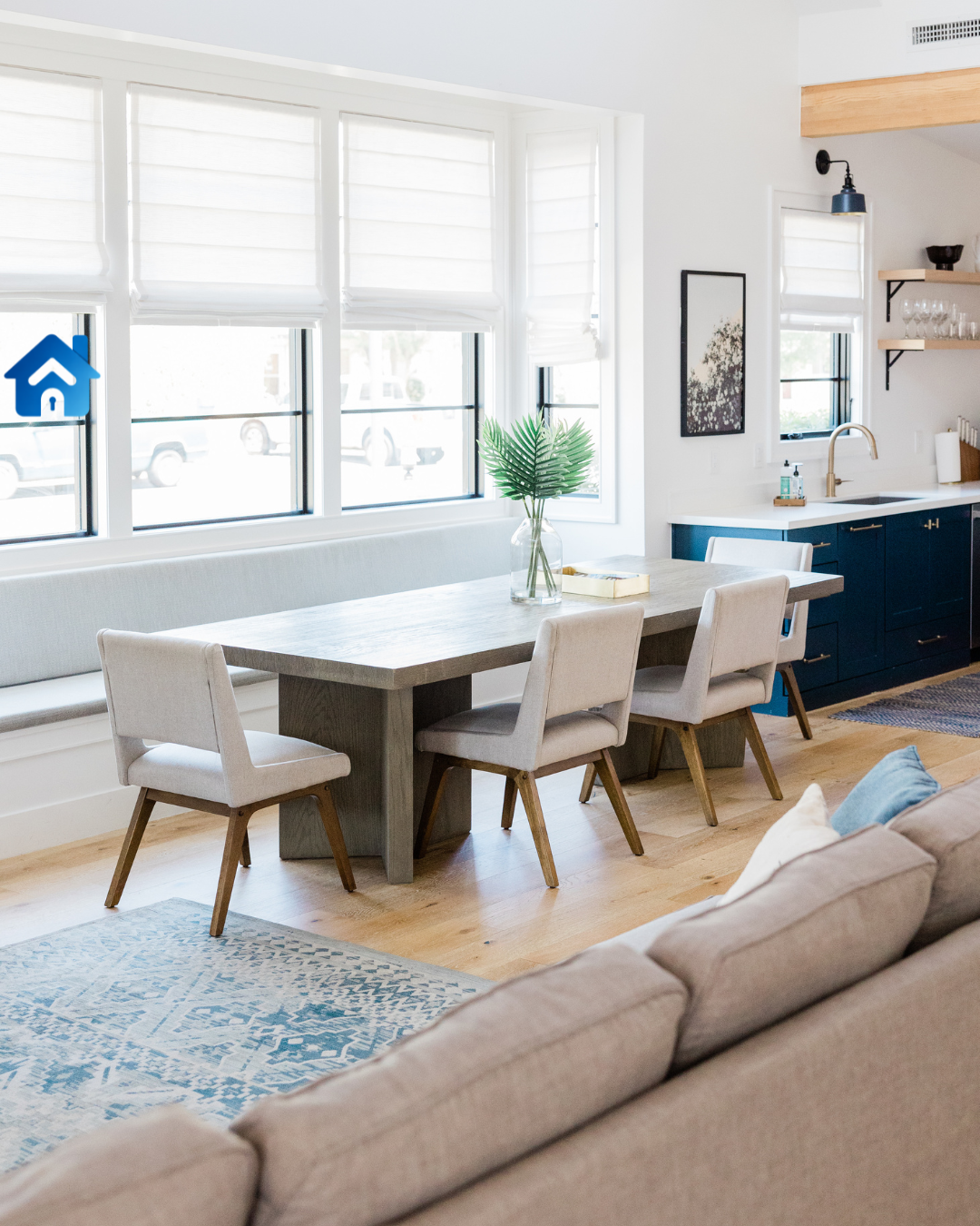 Modern dining area with a wooden table, six upholstered chairs, a glass vase with green leaves, large windows with white blinds, and a glimpse of a kitchen with navy cabinets and open shelves.