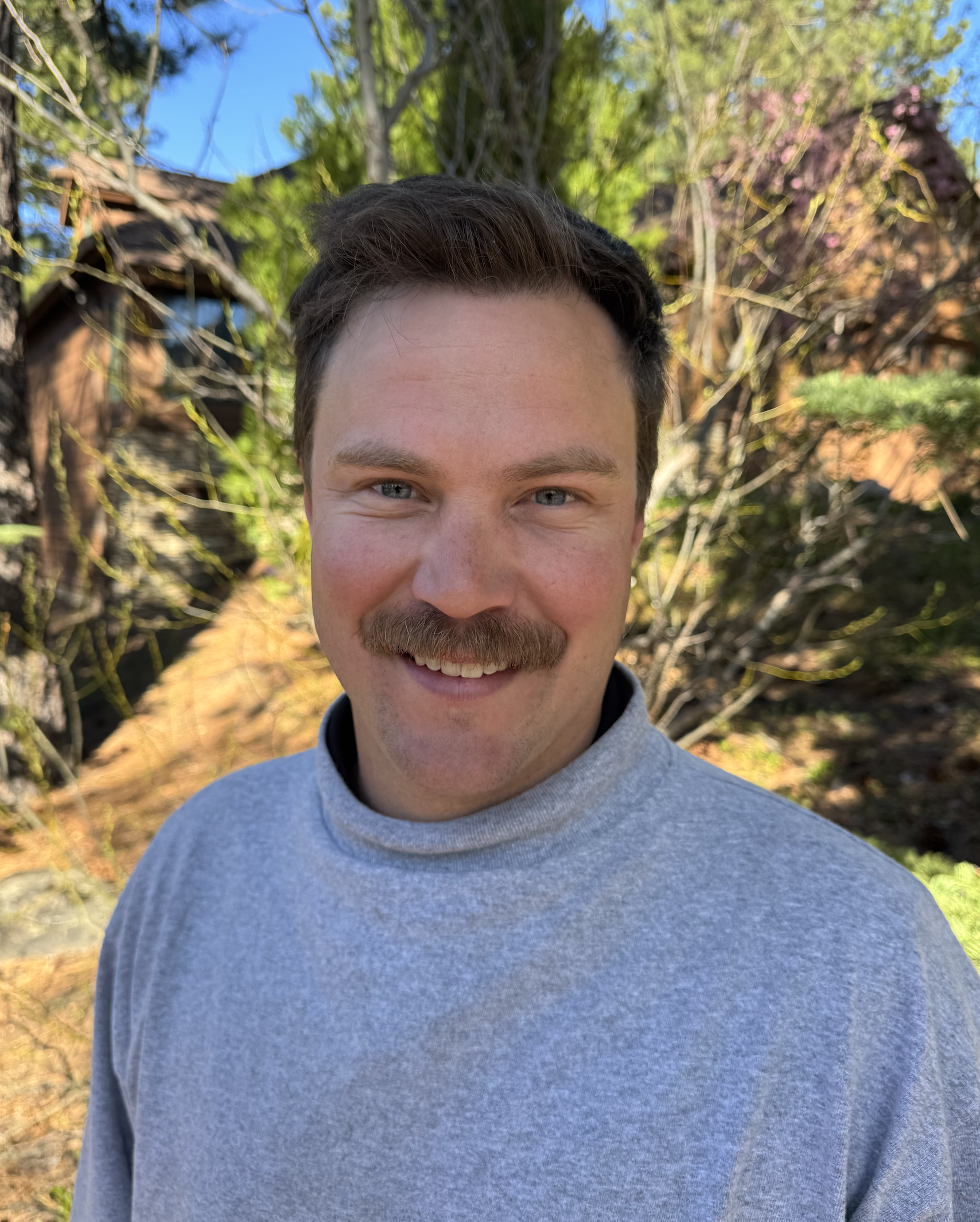 A smiling man with a mustache and short dark hair posing outdoors. He is wearing a light gray turtleneck sweater, with trees and a clear blue sky in the background.