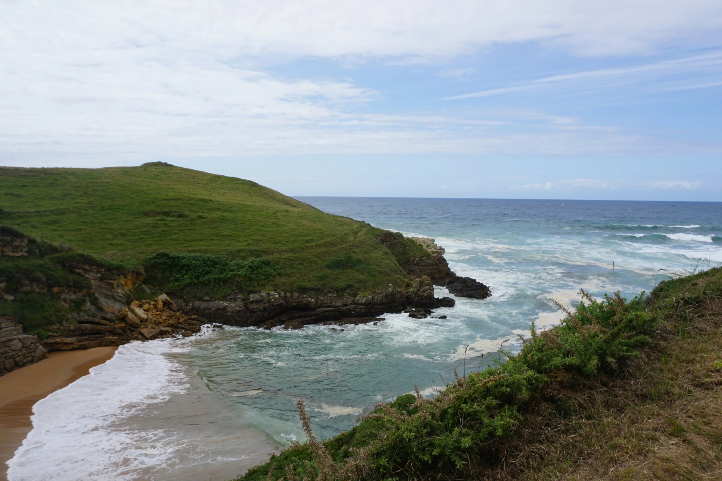 Coastal landscape with green grassy hills, rocky cliffs, sandy beach, ocean waves, and a partly cloudy sky.